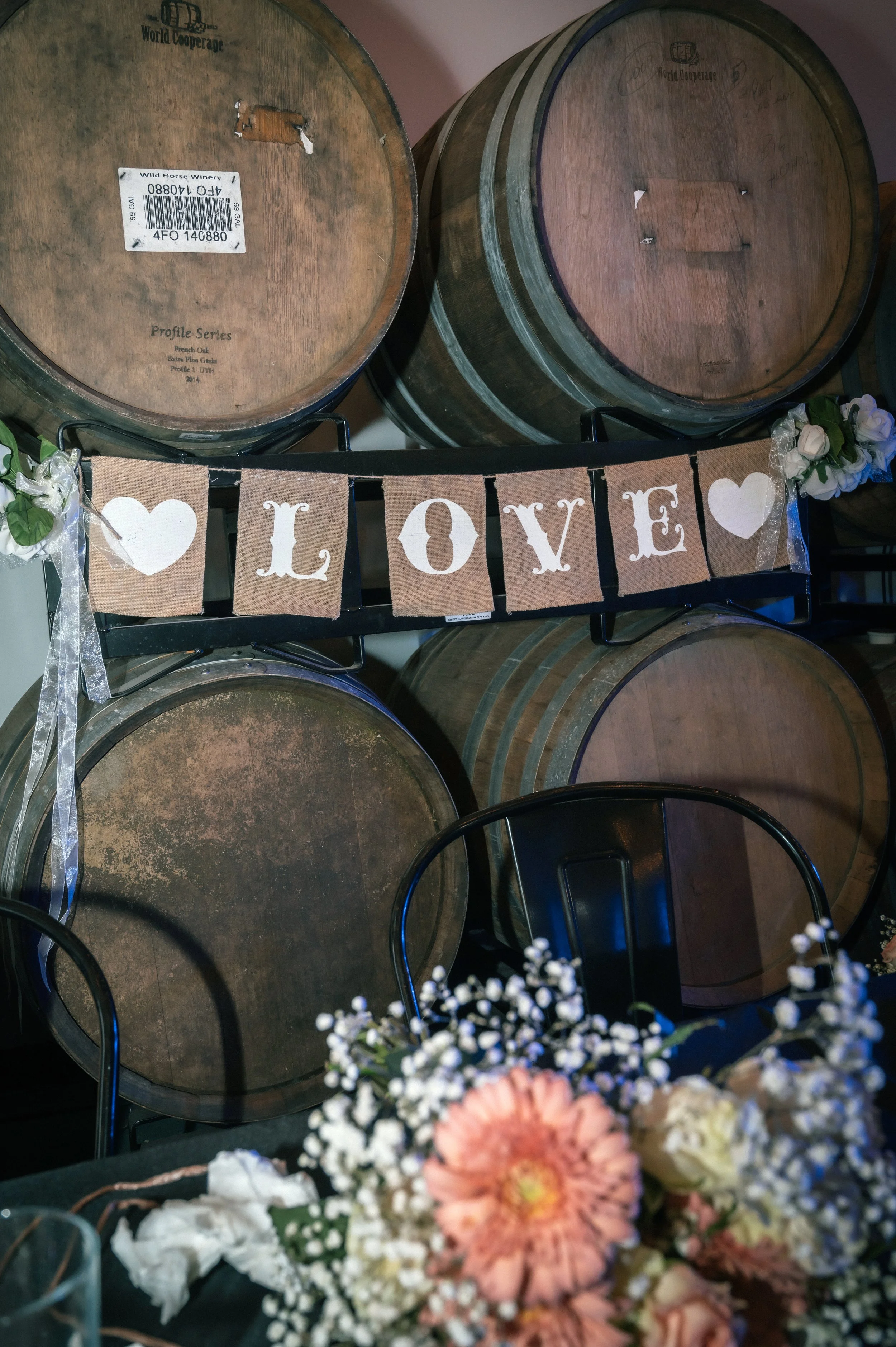 Stacked wooden wine barrels with a burlap banner decorated with white hearts and the word 'LOVE' hanging in front, floral arrangements on the table in front.