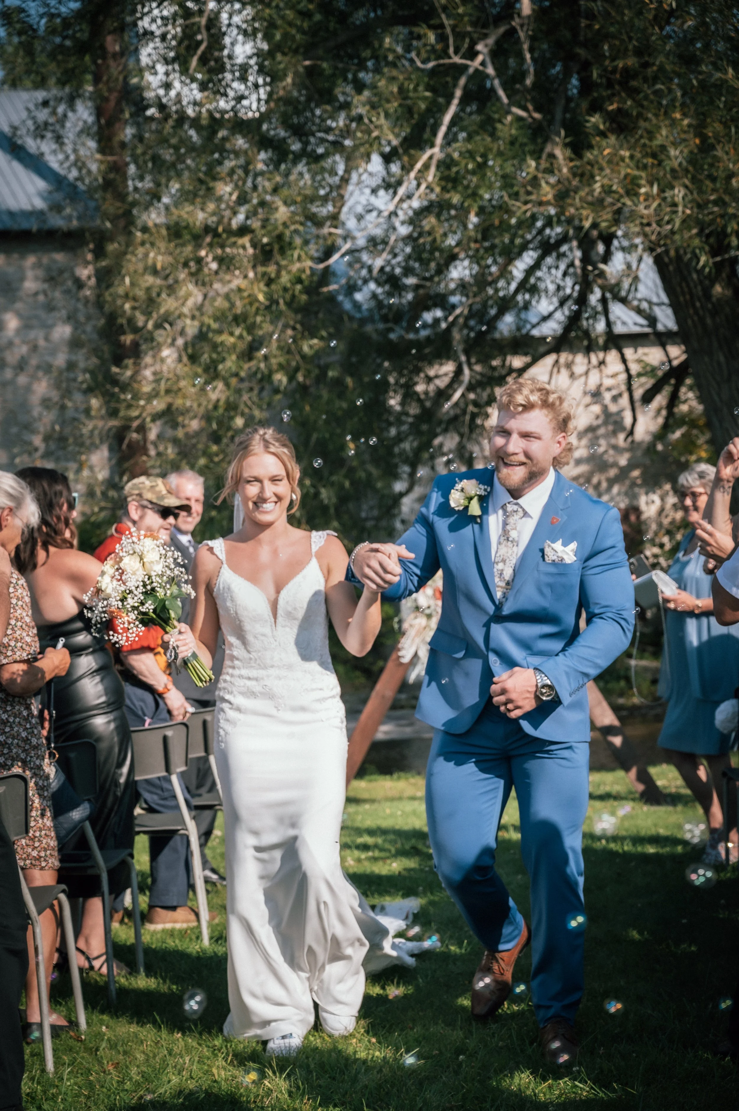Bride and groom walking down the aisle after wedding ceremony, smiling and holding hands, surrounded by wedding guests outdoors.