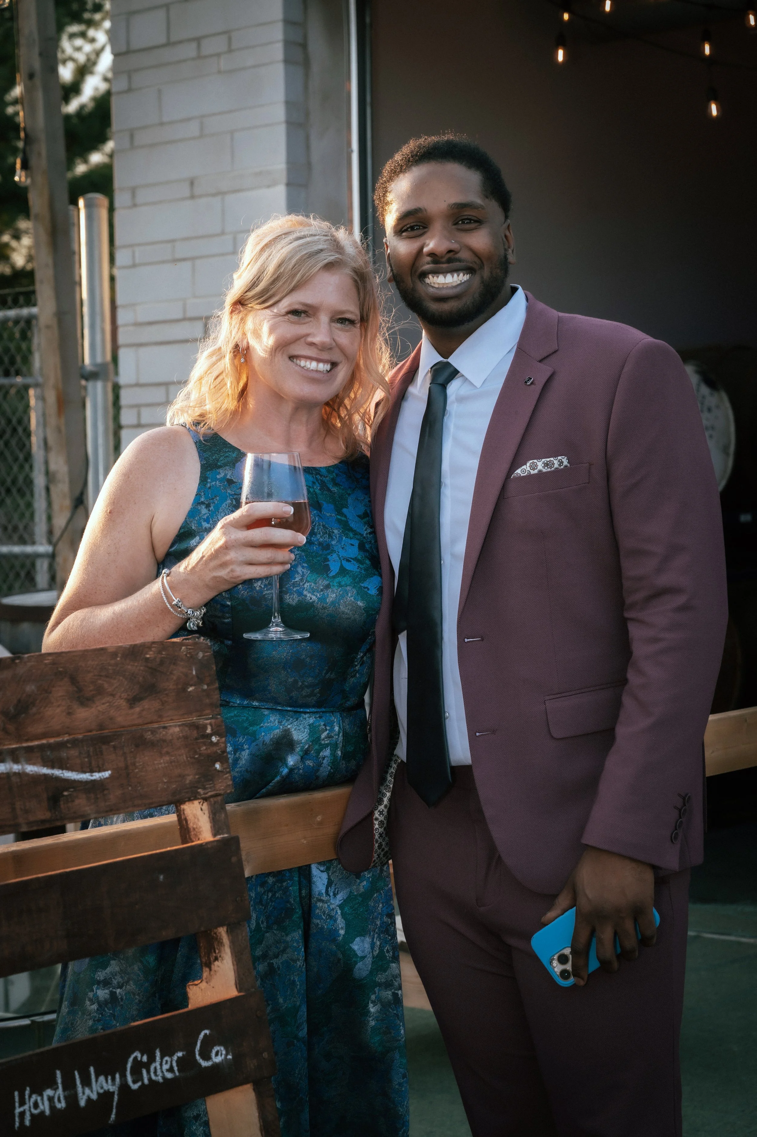 A woman in a blue dress holding a glass of wine and a man in a burgundy suit with a tie, smiling and standing close together at an outdoor event.