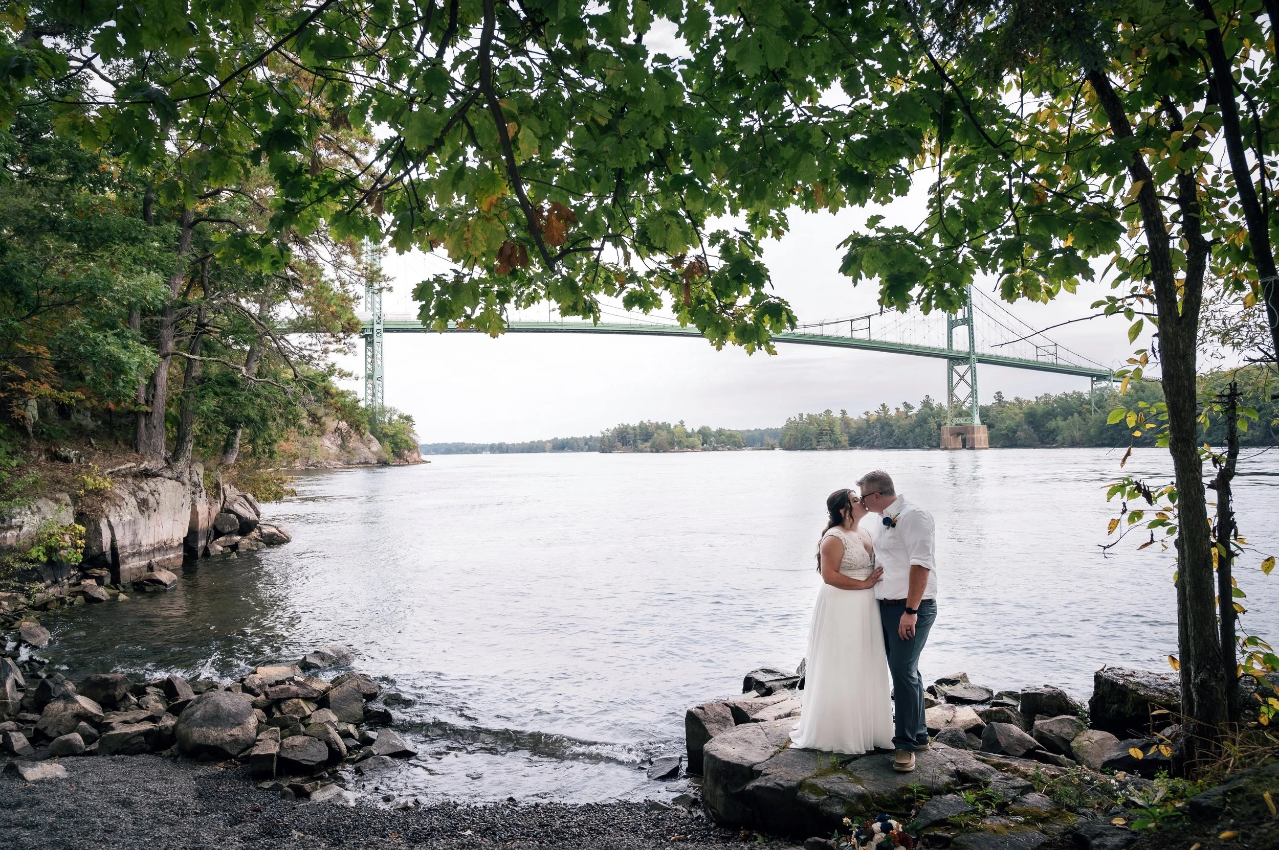 A couple in wedding attire kissing on rocks at a riverside, with a bridge in the background and trees framing the scene.