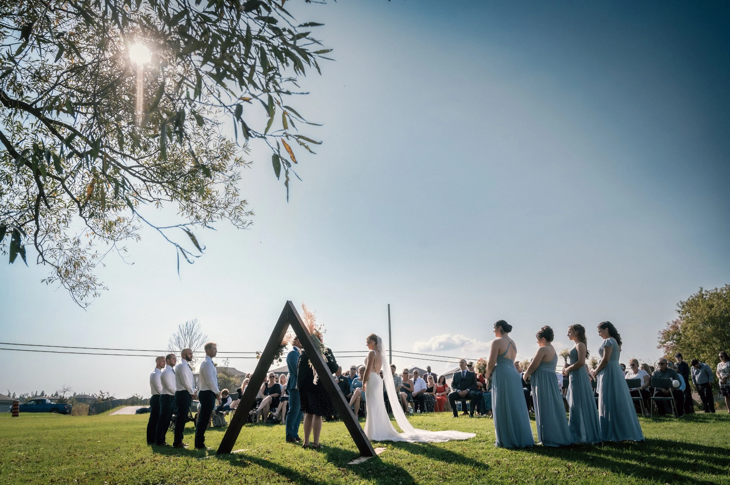 Outdoor wedding ceremony with the bride and groom standing under an arched structure, surrounded by bridesmaids in blue dresses, officiant, and seated guests under a clear sky with sunlight filtering through tree branches.