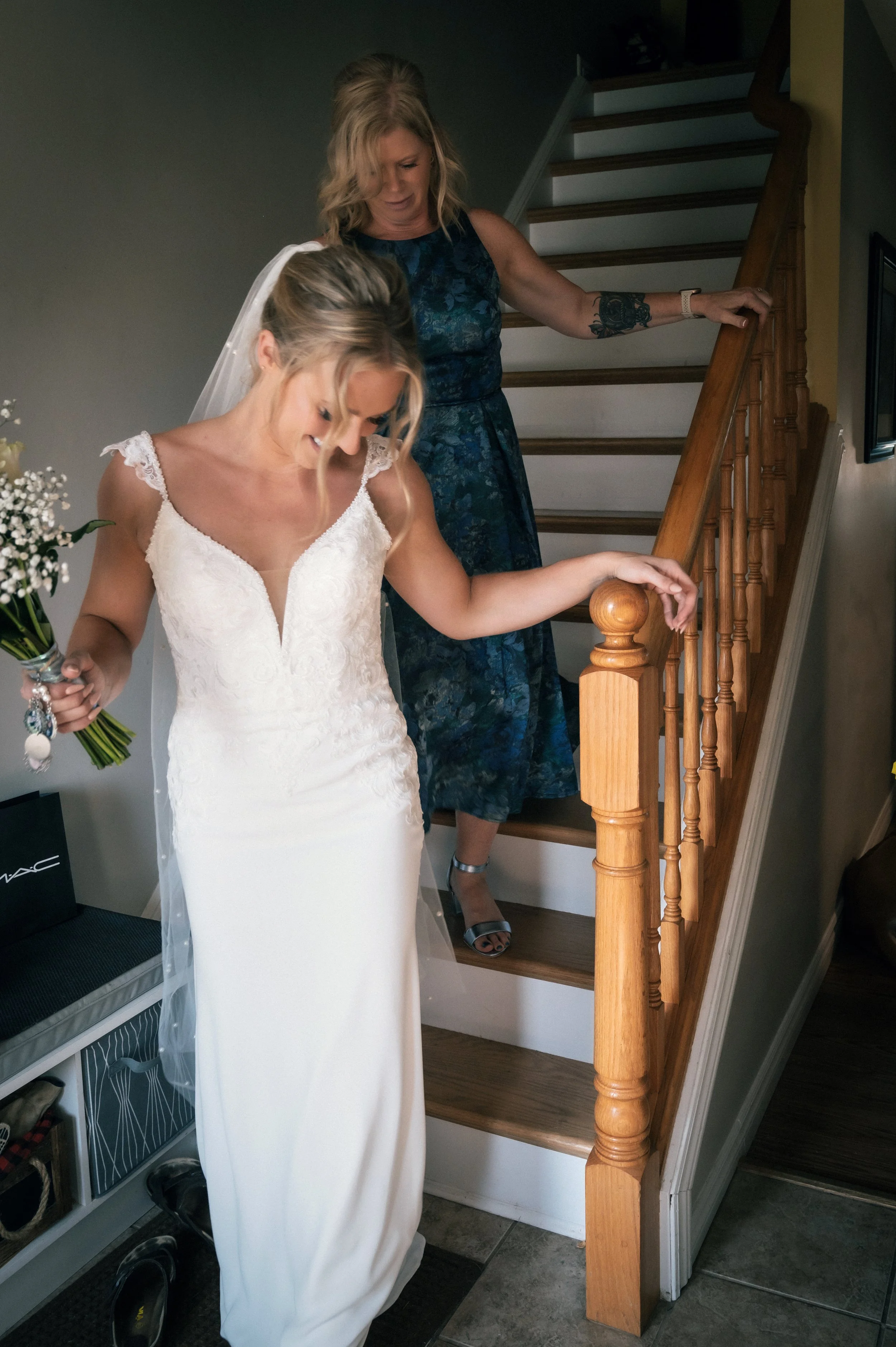 A bride in a white wedding gown with a veil, holding a bouquet, descending stairs with a woman in a blue dress.