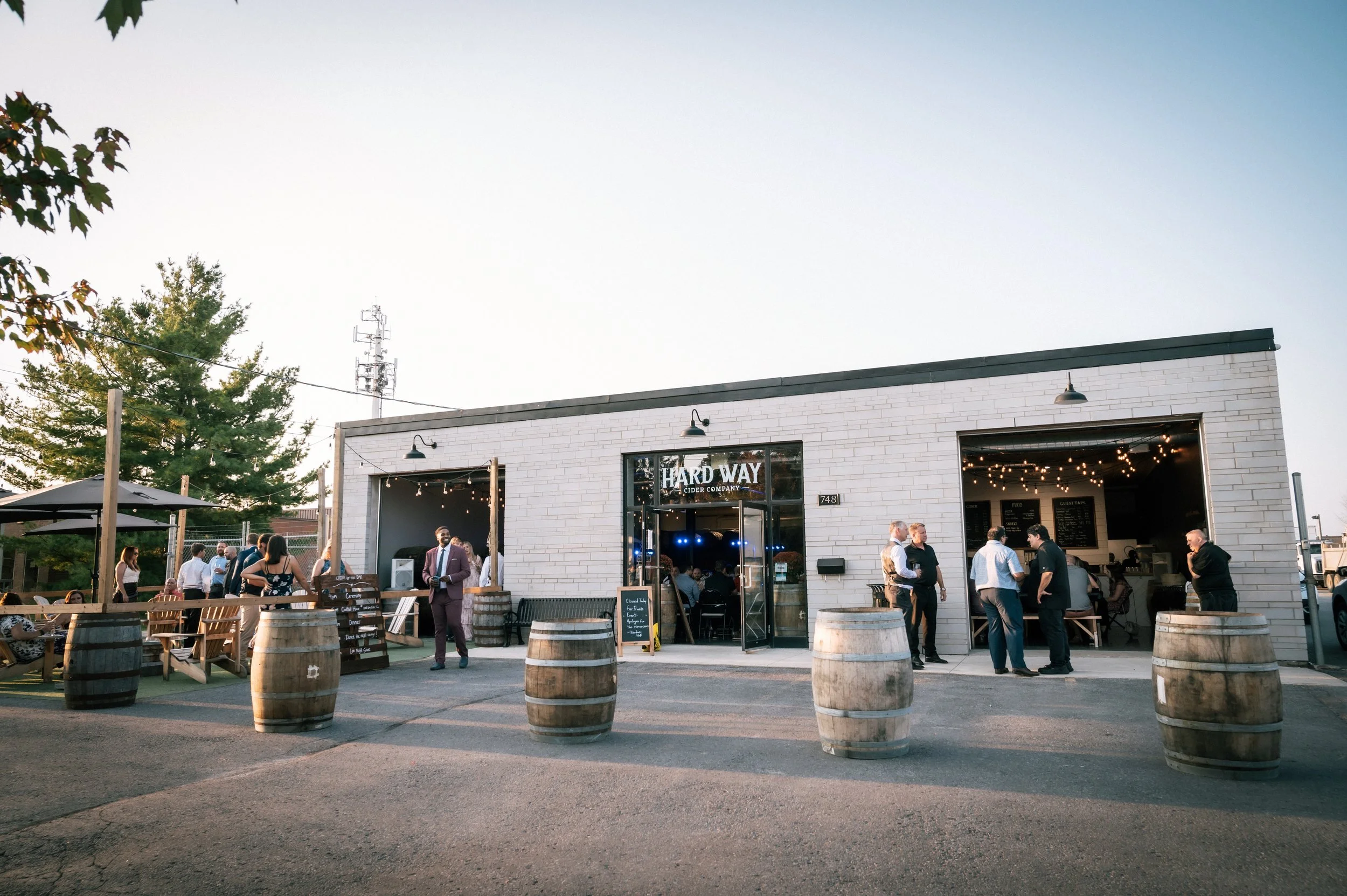 People socializing outside of a brewery named 'Hard Way Cider Company' in a modern, white brick building with large open windows decorated with string lights. Wooden barrels are placed around the outdoor seating area.