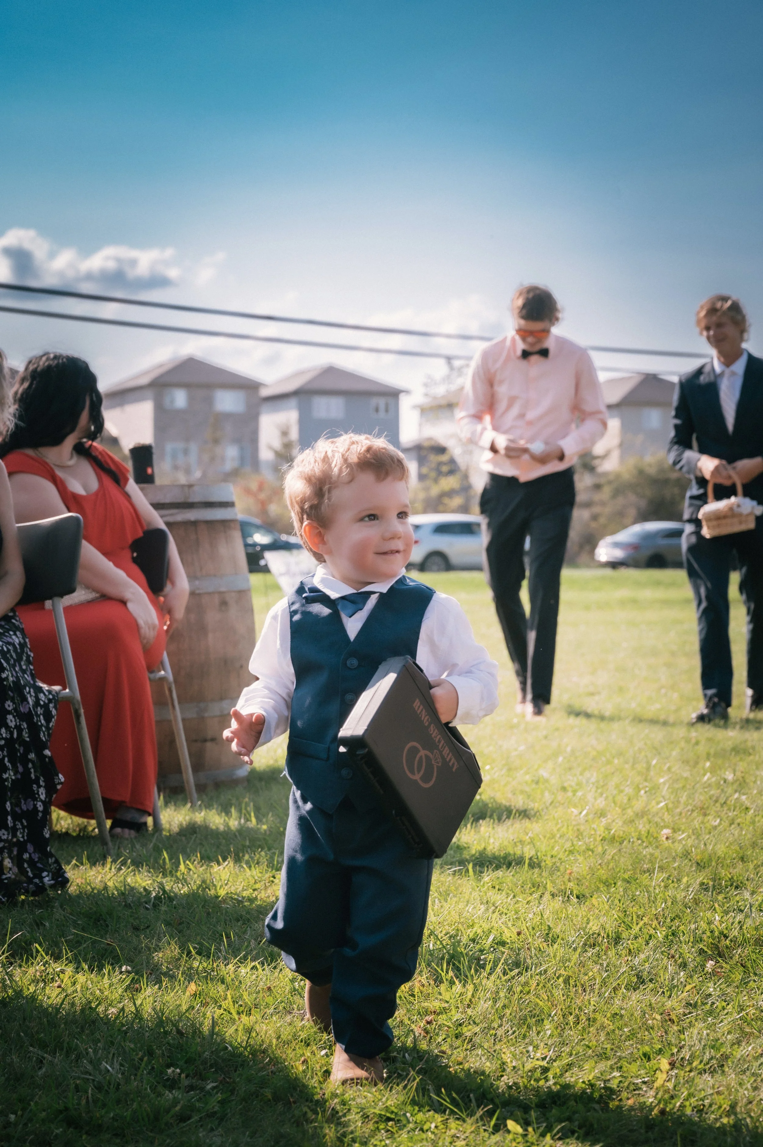 A young boy in a navy vest and white shirt walking barefoot outside during a sunny daytime event, carrying a briefcase labeled 'KING SECURITY,' with several adults in formal attire and houses in the background.