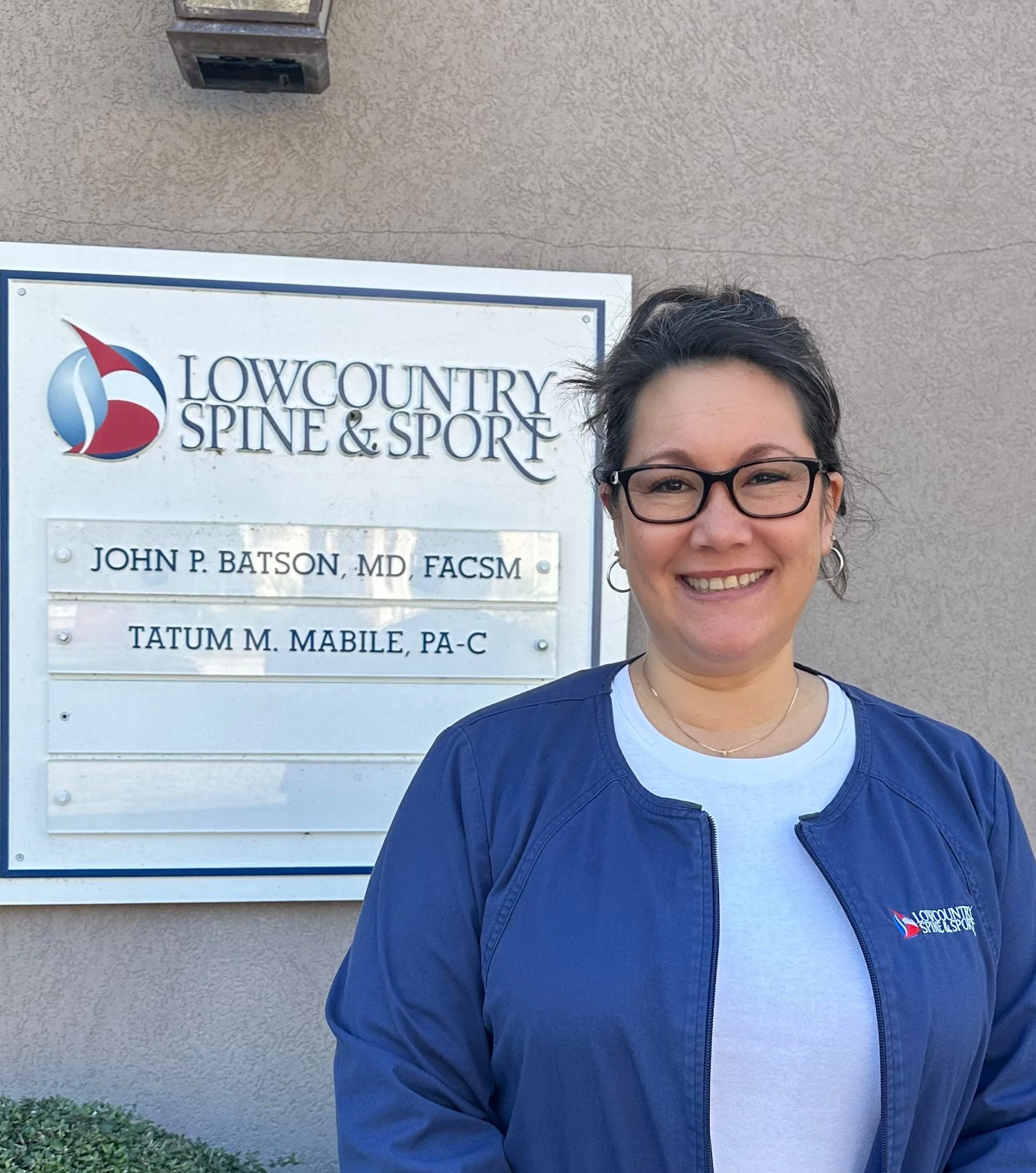 A woman standing outside Lowcountry Spine & Sport clinic, smiling, wearing glasses and a blue jacket with the clinic's logo. There is a sign with the clinic's name and doctors' names next to her.