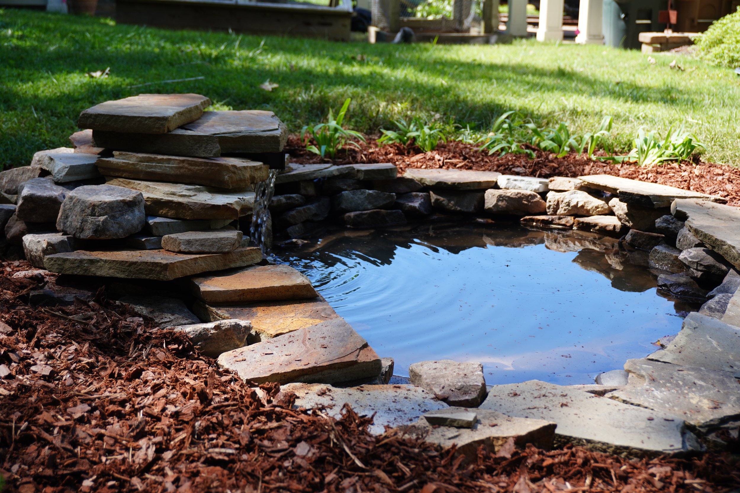 A small backyard pond surrounded by stacked flat stones and rocks, with a small waterfall feature at one end, in a grassy yard with green plants and a wooden deck in the background.