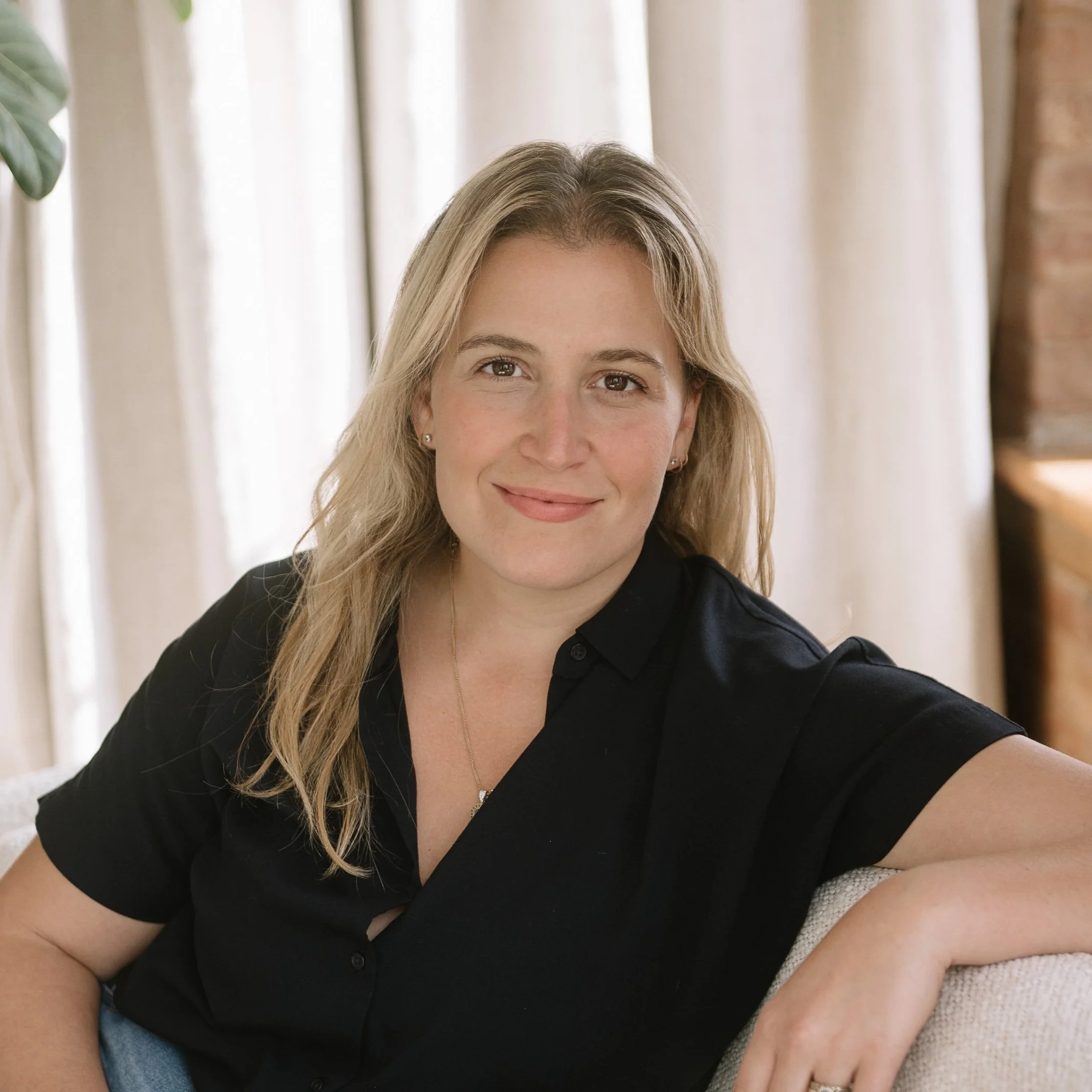 A woman with long blonde hair and a gentle smile, wearing a black shirt, sitting on a light-colored couch in a room with beige curtains and a brick wall in the background.
