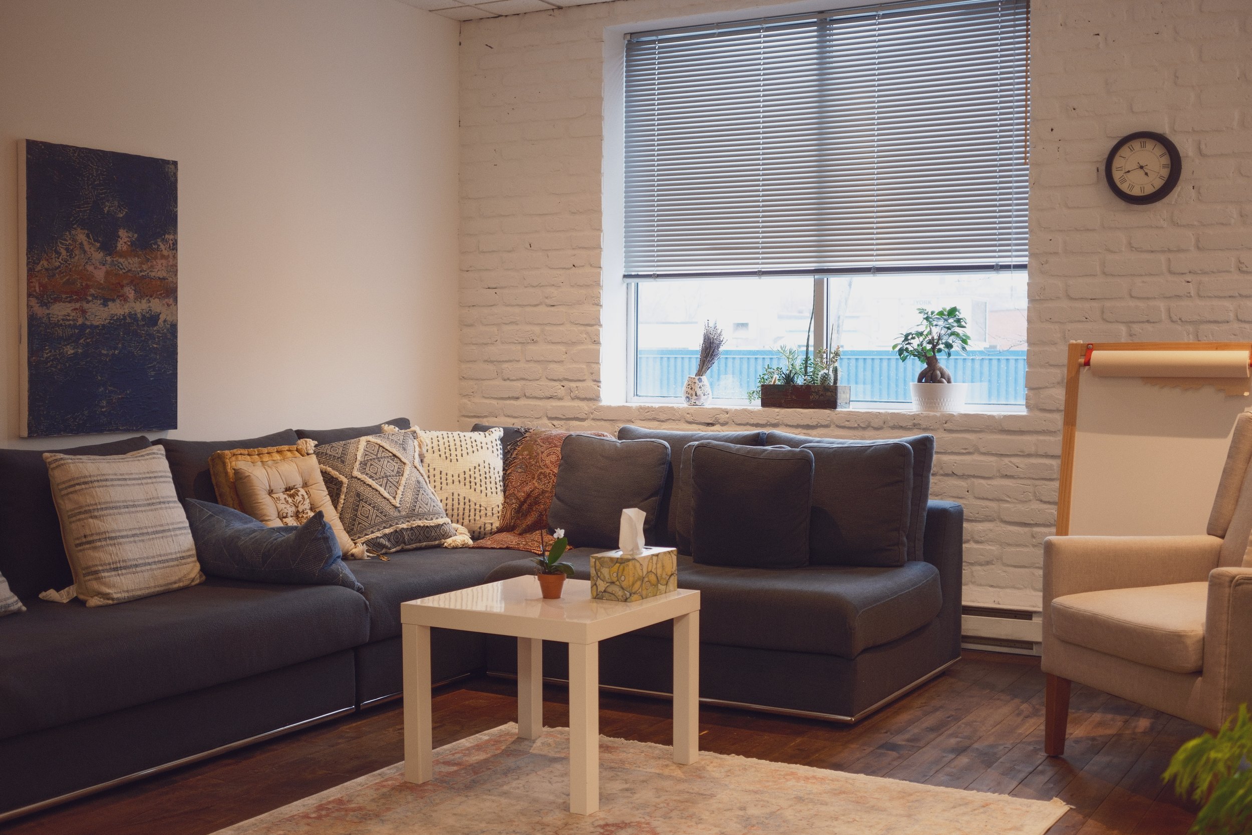 Office 2: This large and thoughtfully designed office features a comfortable sectional sofa and armchair, ideal for group or individual sessions. Natural light fills the space through large windows, complemented by lush greenery. A charming dollhous