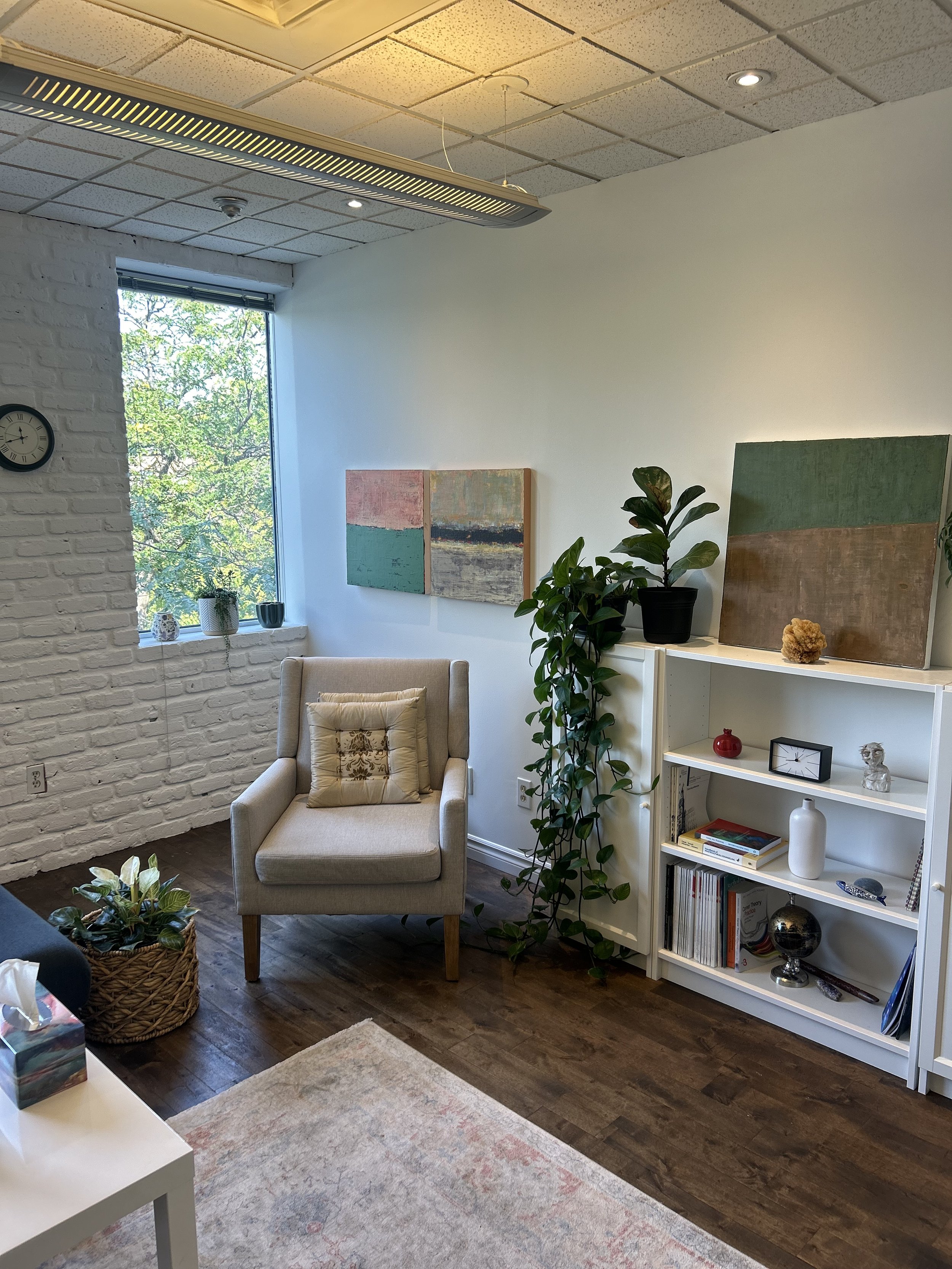 Office 2: This large and thoughtfully designed office features a comfortable sectional sofa and armchair, ideal for group or individual sessions. Natural light fills the space through large windows, complemented by lush greenery. A charming dollhous