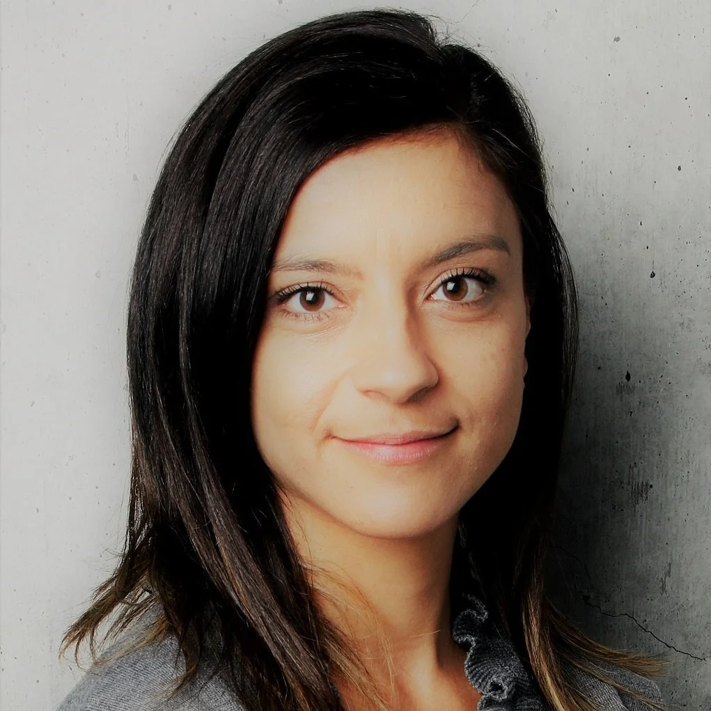 Close-up portrait of a woman with brown eyes and dark brown hair smiling softly, wearing a gray top, against a neutral gray wall background.