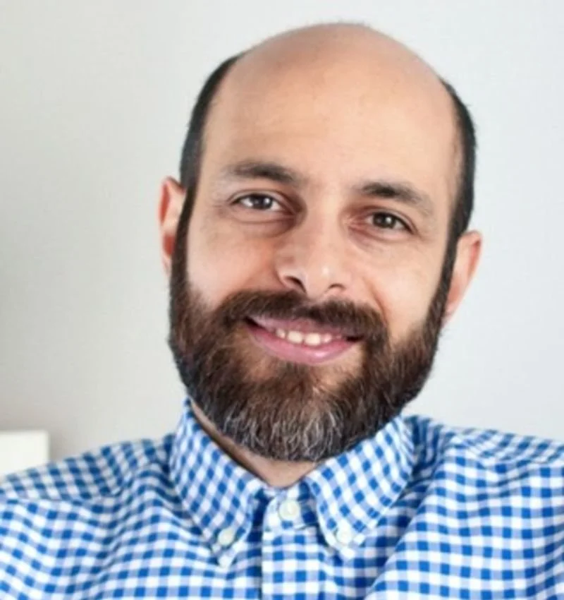 A man with a beard and mustache, wearing a blue and white checkered shirt, smiling at the camera.