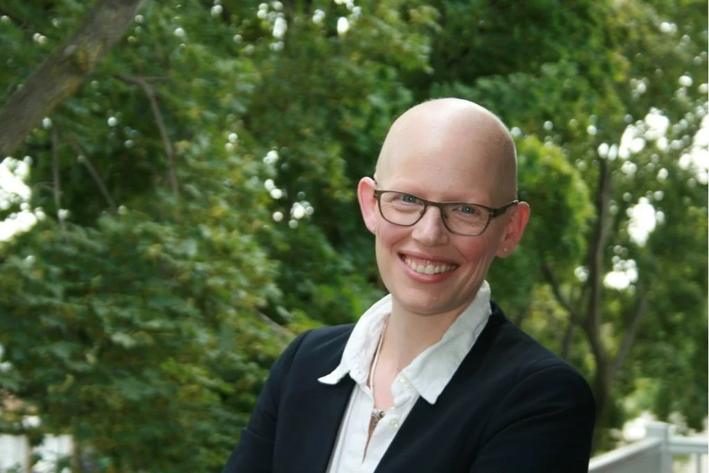 Smiling woman with glasses and a shaved head standing outdoors with greenery in the background.