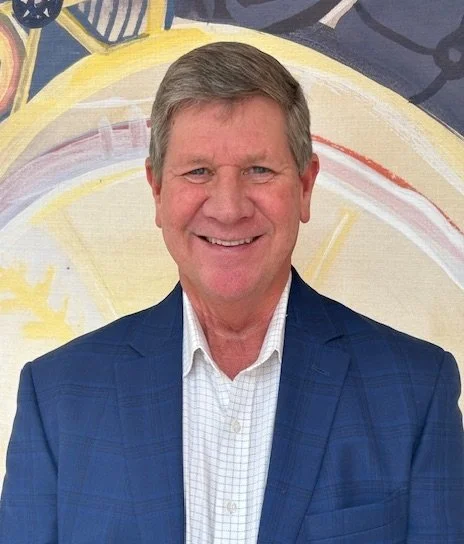 A smiling man in a blue suit and white shirt standing in front of a colorful, abstract background.