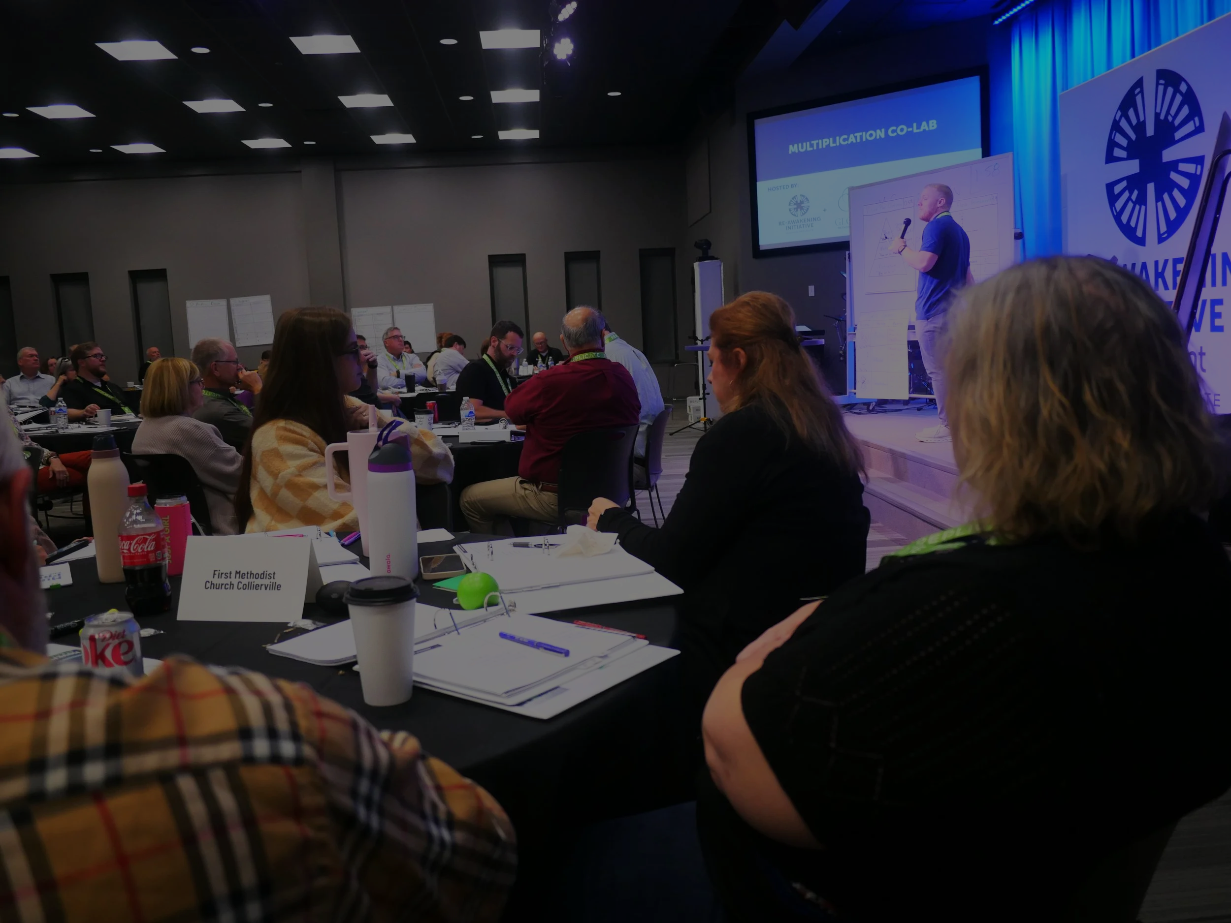People attending a conference in a large room, listening to a speaker on stage with a presentation slide, at a conference titled 'Multiplication Co-Lab'.
