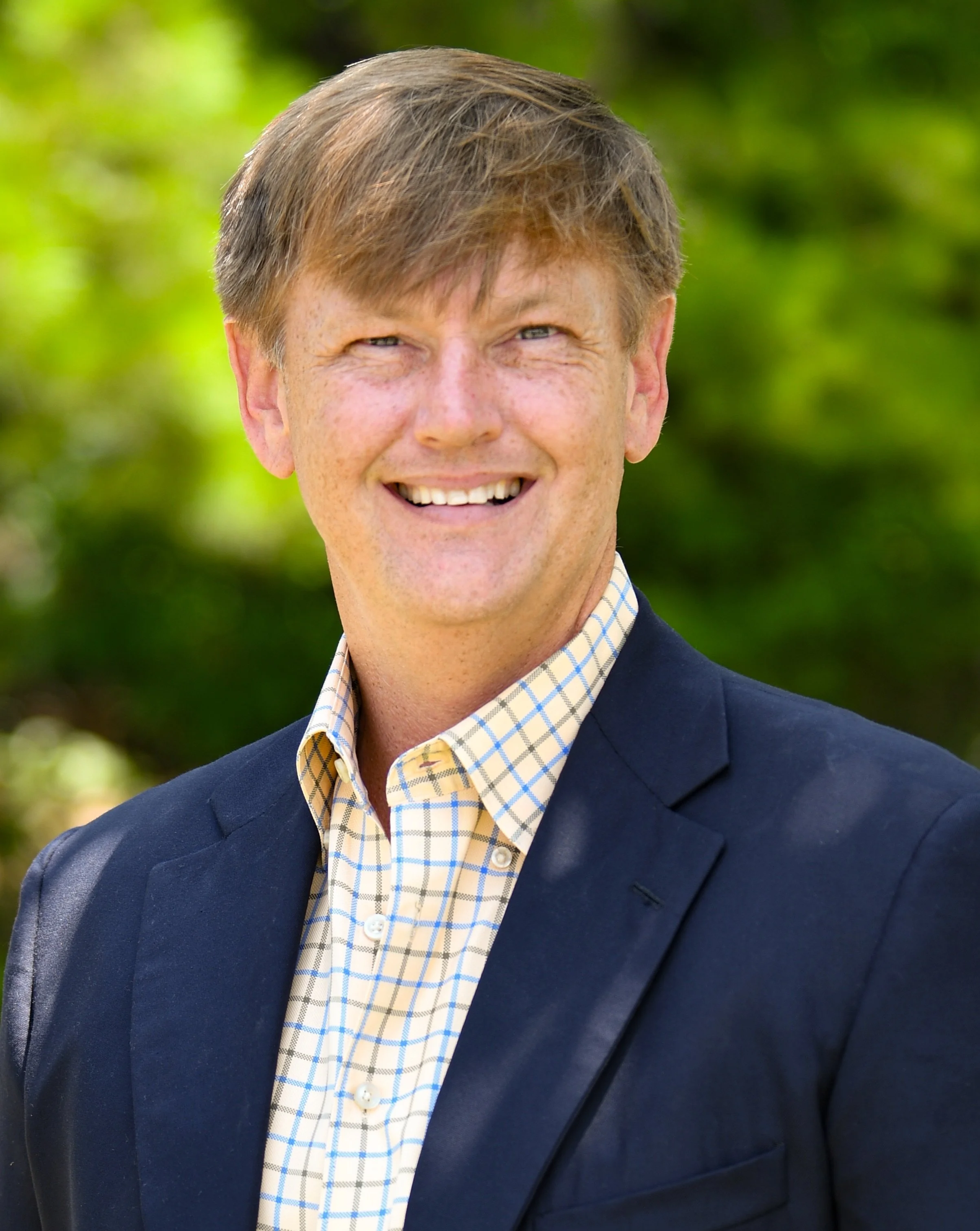 A smiling man with light brown hair wearing a navy blazer and a yellow checkered shirt outdoors with green foliage in the background.