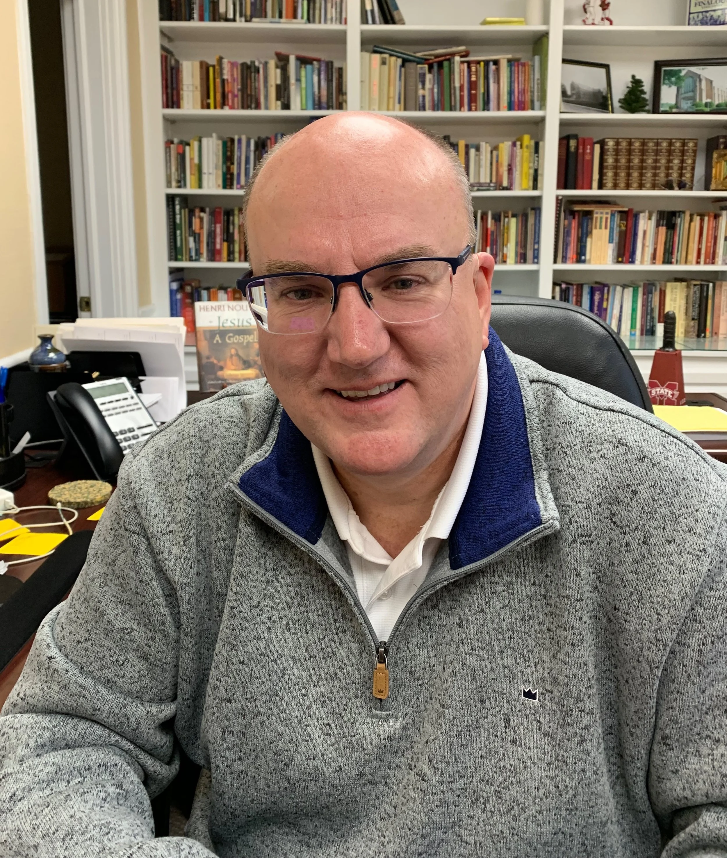A smiling man with glasses sitting at a desk in an office with a bookshelf filled with books in the background.
