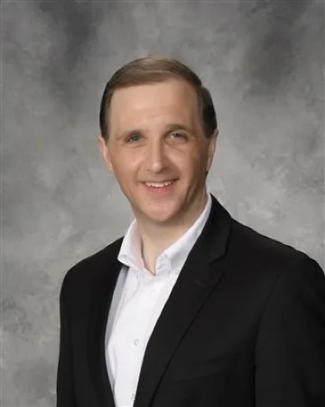A man with light brown hair wearing a black suit jacket and white shirt, smiling against a gray background.