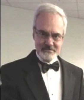 A man with gray hair and glasses wearing a tuxedo and bow tie, standing indoors.