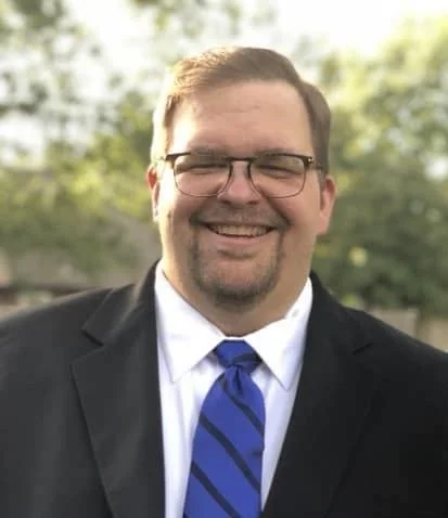 Man in suit smiling outdoors with trees in the background.
