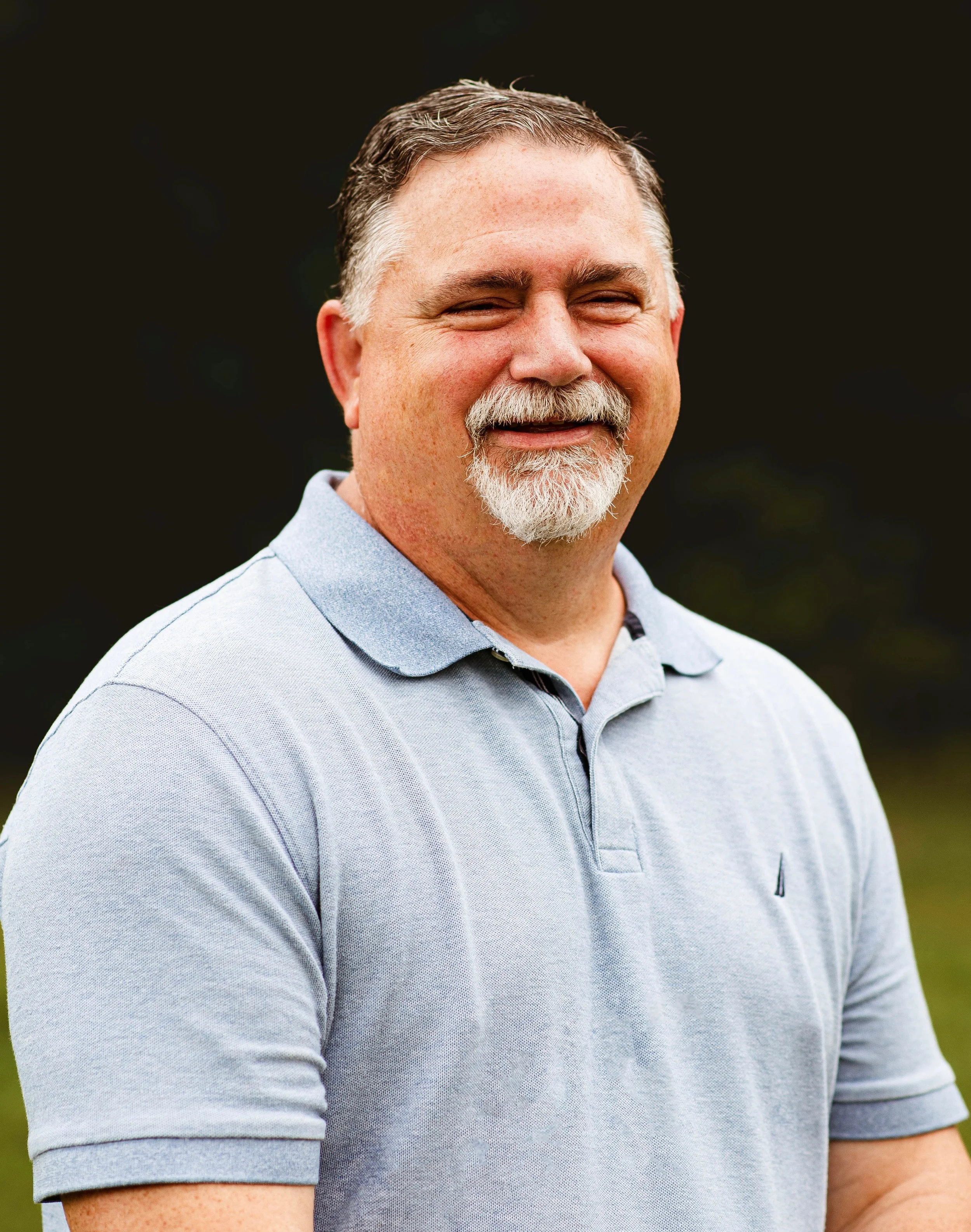 A smiling middle-aged man with a beard and mustache wearing a light gray polo shirt standing outdoors against a dark blurred background.