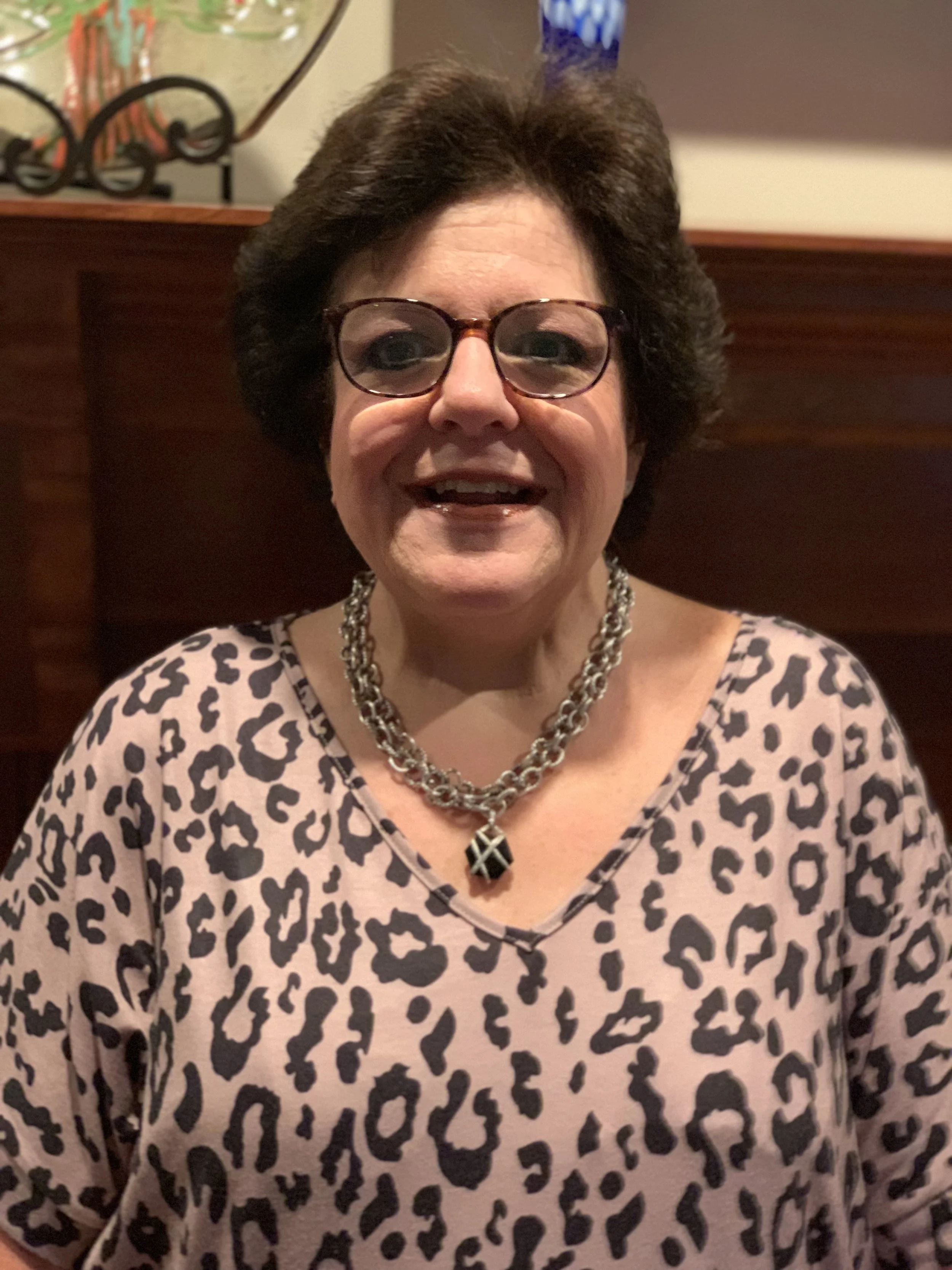 Smiling woman with short brown hair, glasses, and a leopard print top, wearing a chunky silver necklace with a pendant, in a warmly lit room.