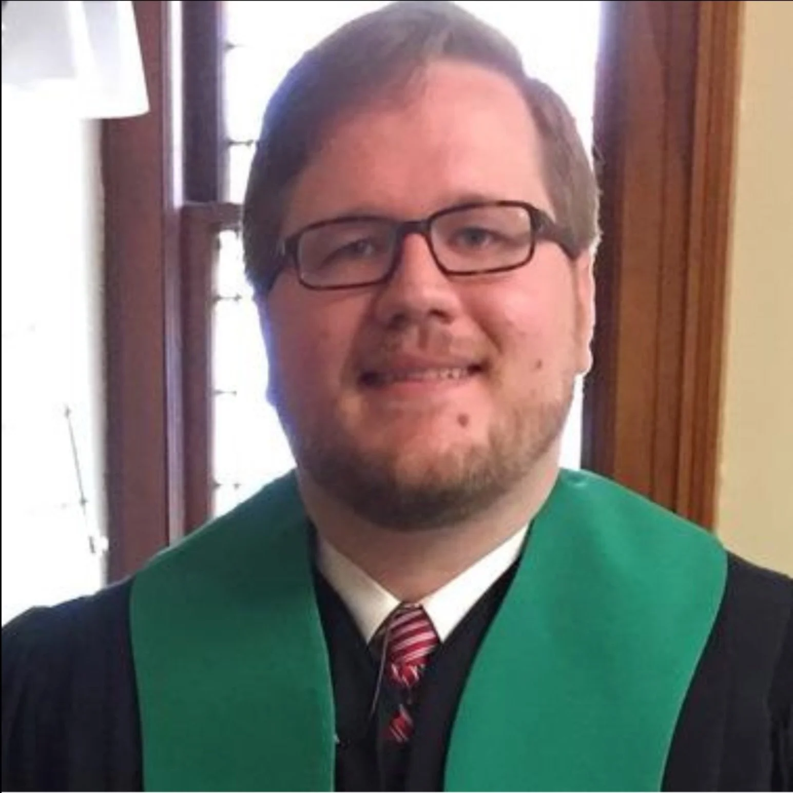 A man in a black graduation gown and green stole, wearing glasses, with short brown hair and a beard, smiling, standing indoors near a window with wooden trim.