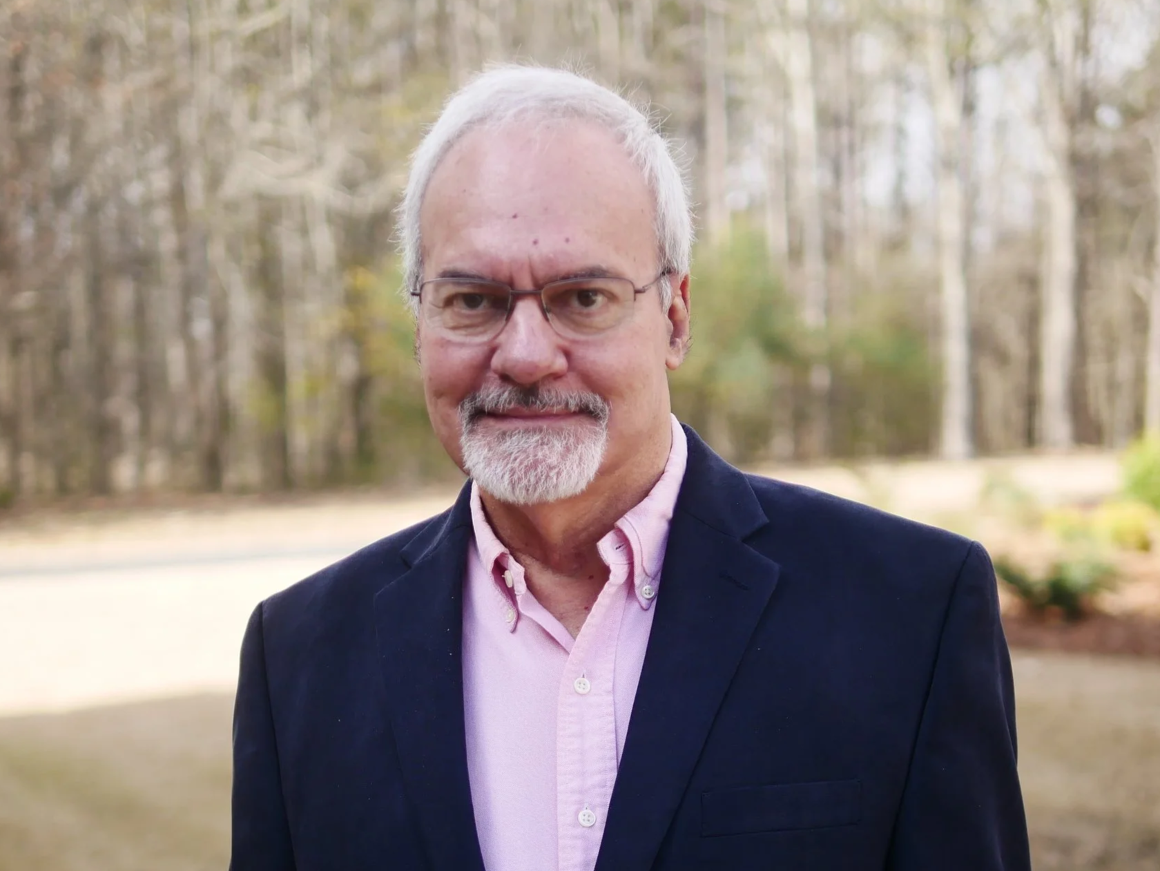 A middle-aged man with white hair, glasses, and a beard smiling outdoors. He is wearing a pink button-up shirt and a dark blazer, with a background of trees.