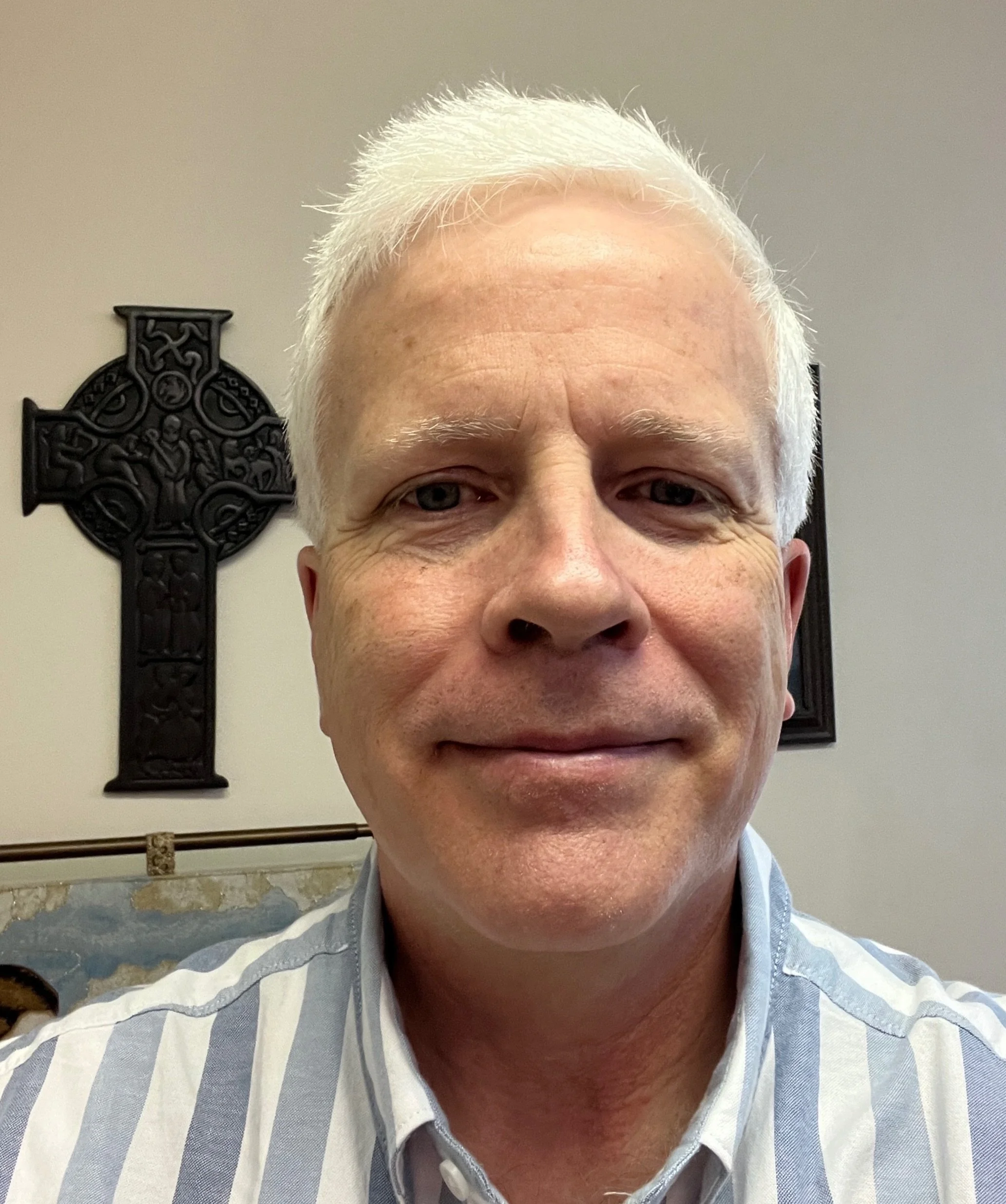 A close-up portrait of a smiling middle-aged man with white hair, wearing a striped shirt, standing indoors with a decorative black cross on the wall behind him.