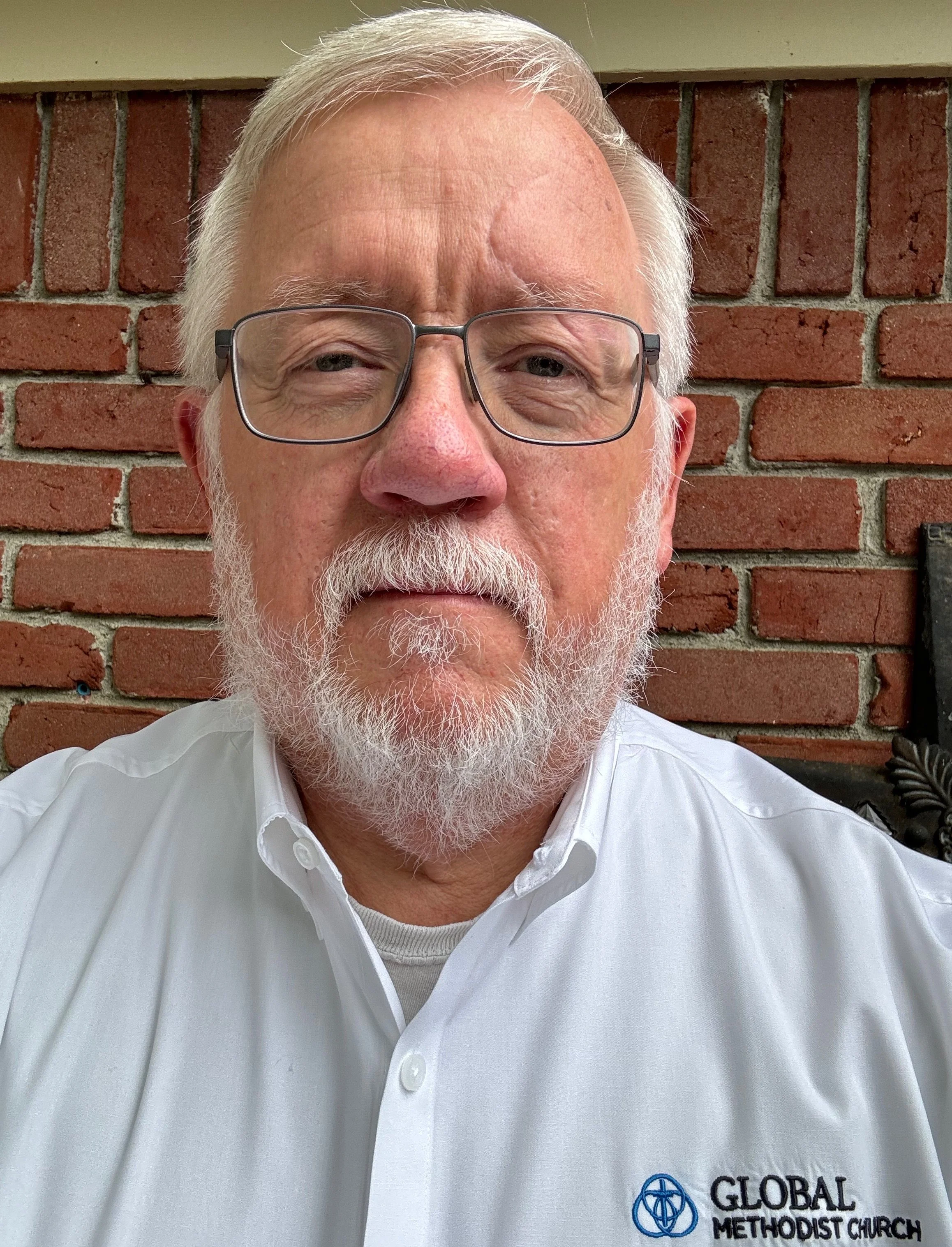 A middle-aged man with glasses, white hair, and a beard wearing a white shirt with a 'Global Methodist Church' logo, standing in front of a brick wall.