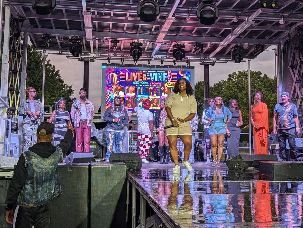 A performer holding a microphone stands in front of a group on the Live on the Vine stage.