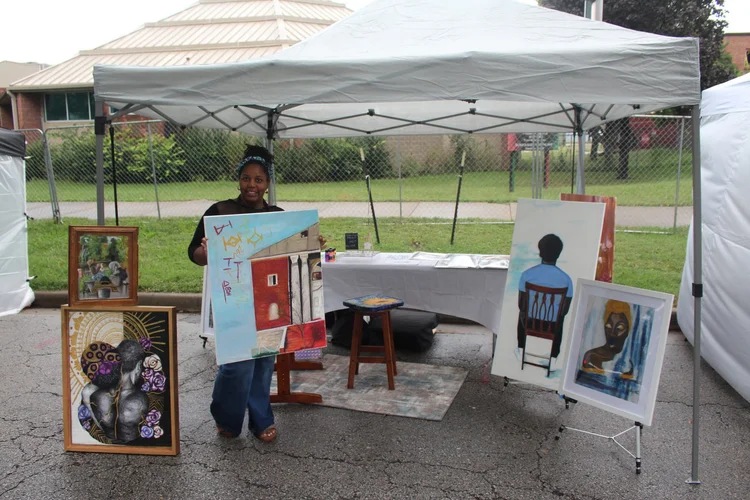 A participant holding artwork beside a booth display. 18th & Vine Arts Festival 2026.