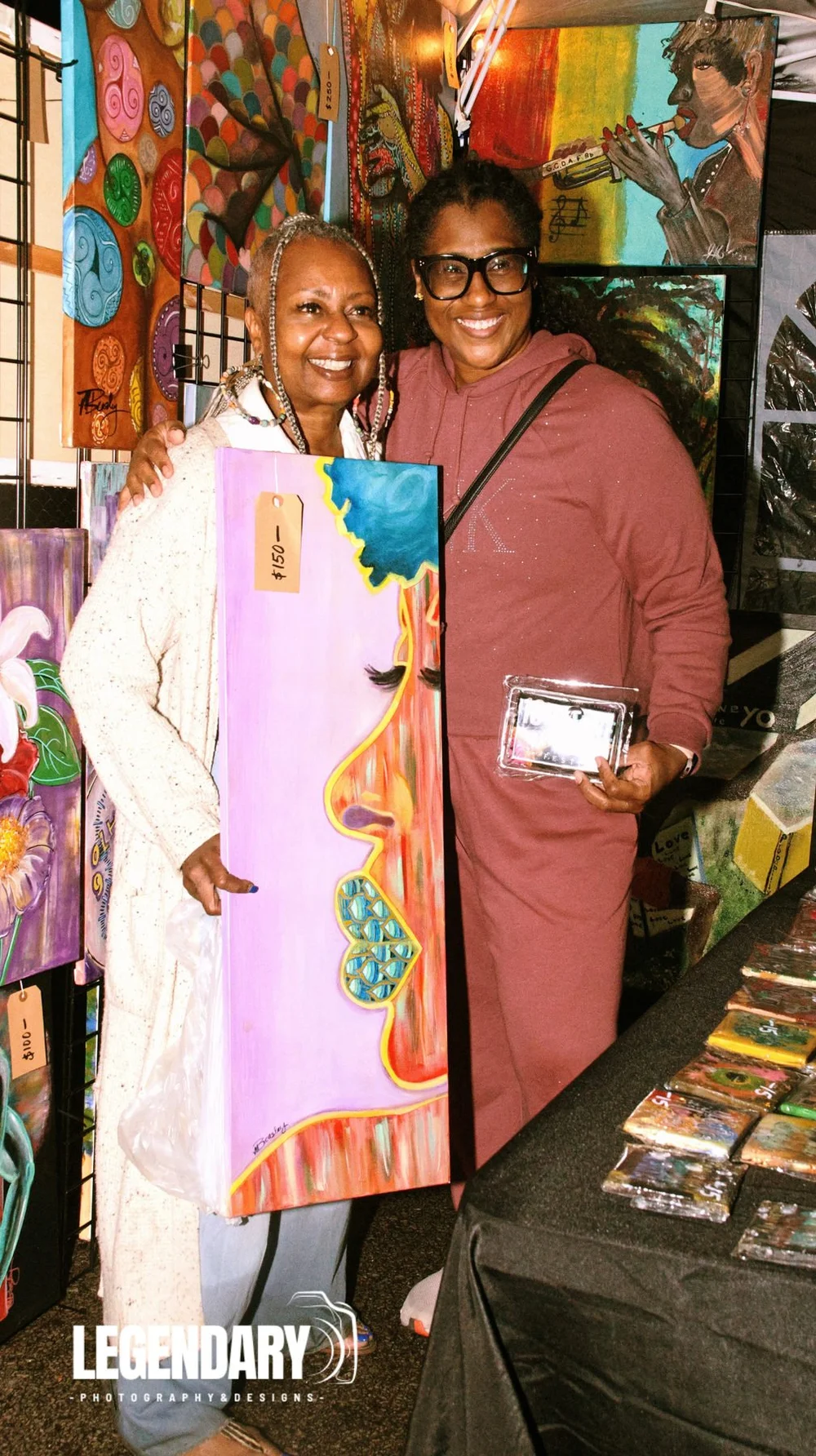 Two smiling participants hold a colorful portrait painting beside other displayed artwork.