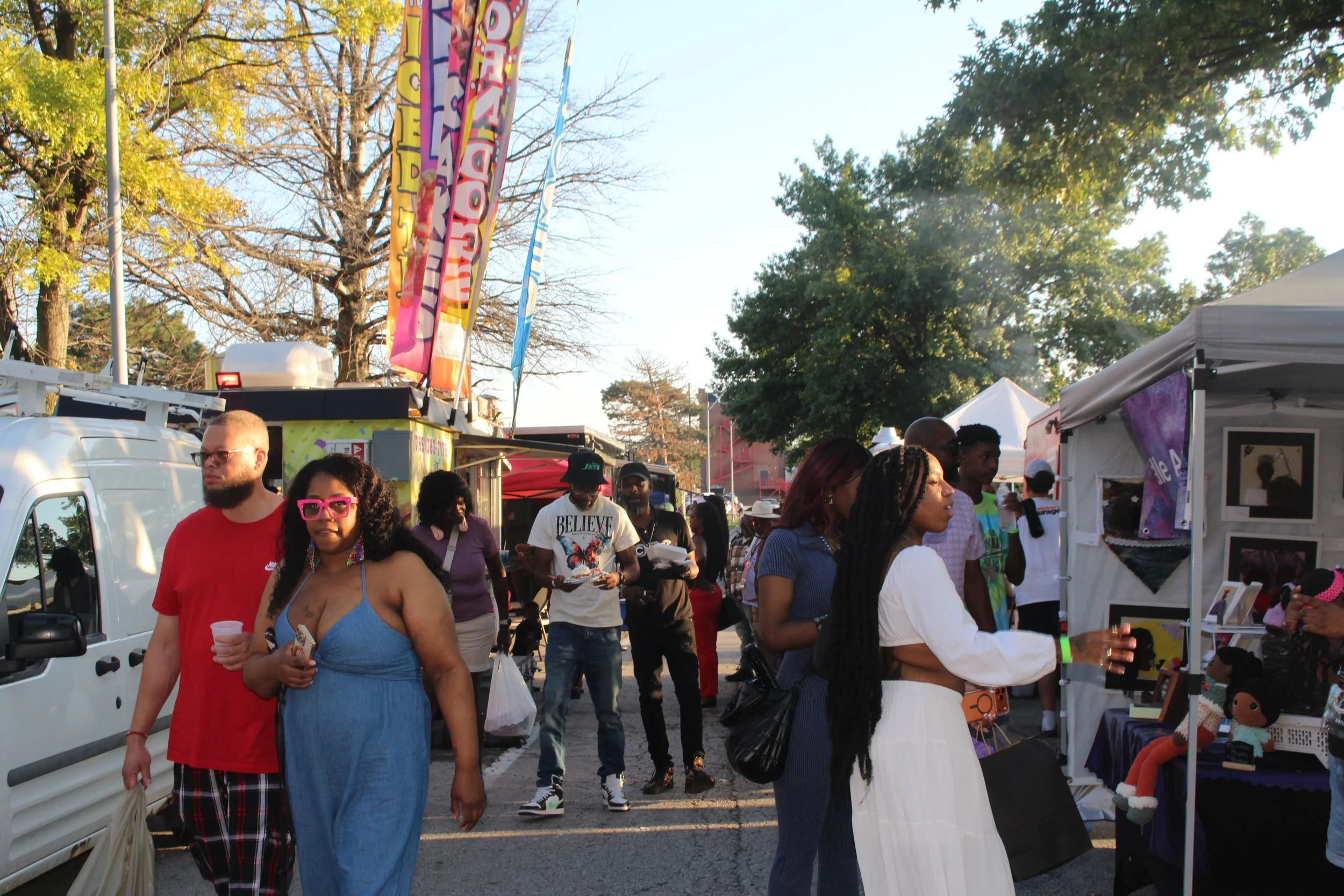 Crowd of people at an outdoor fair or festival with vendor tents, flags, and trees in the background. People are walking and browsing stalls.