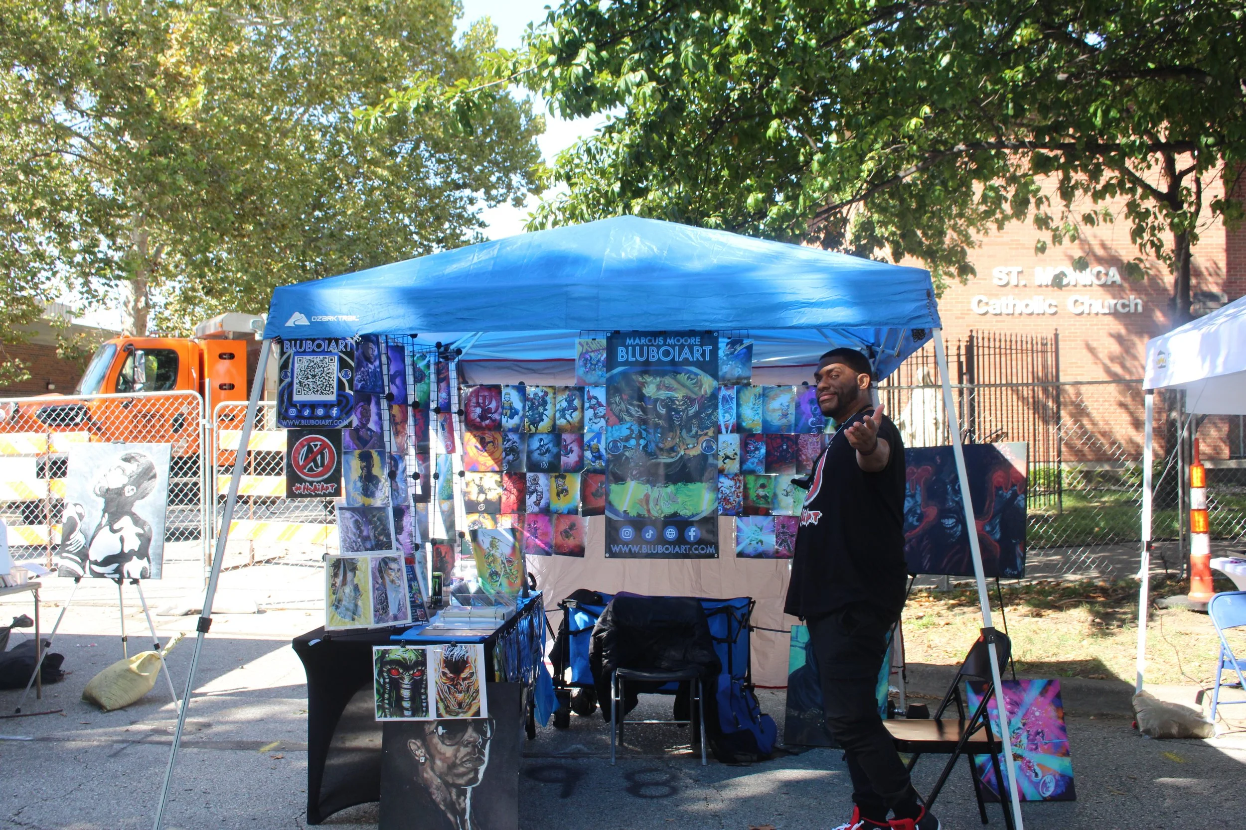 An outdoor art booth with colorful artwork displayed under a blue canopy, with a man standing and gesture towards the artist. In the background, there are trees, a brick building with a sign that reads "St. Monica Catholic Church," and a chain-link fence.