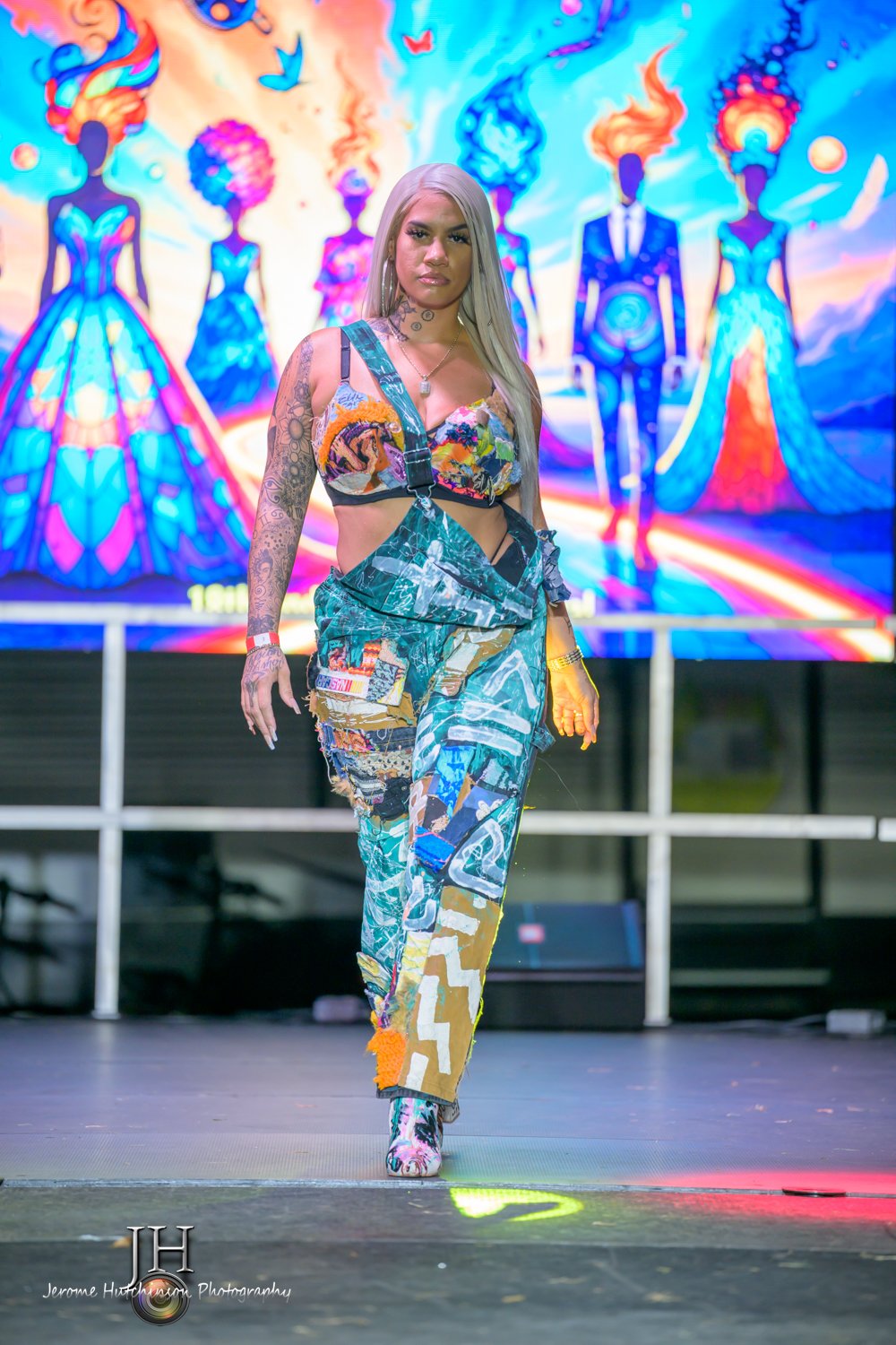 A woman with long gray hair and tattoos on her arms walks on a stage with a colorful, abstract art background featuring five figures in elaborate gowns and suits with painted or fiery hair.