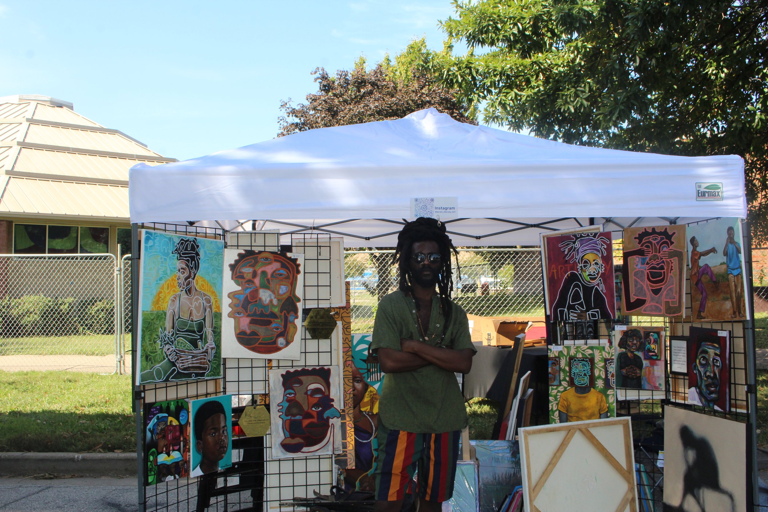 An artist with dreadlocks and sunglasses stands with arms crossed in front of a display of colorful portraits at an outdoor art fair. A white canopy tent shades the artwork, with trees and a building in the background.
