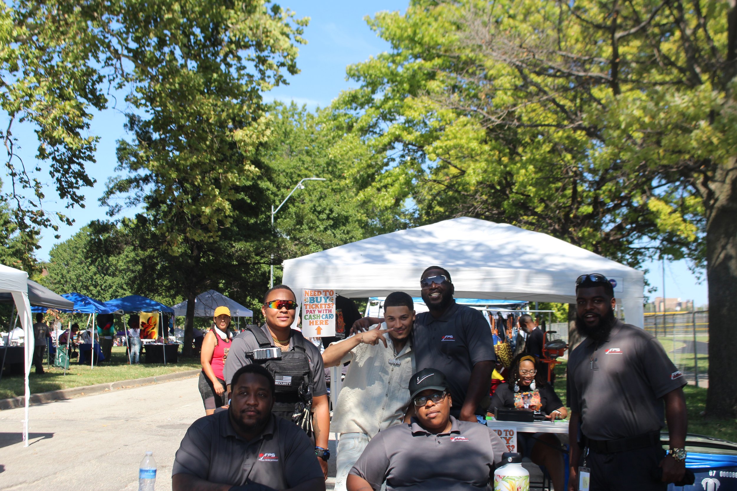 Group of people at an outdoor event under trees, with tents and vendors in the background. Some are smiling and posing for the photo.