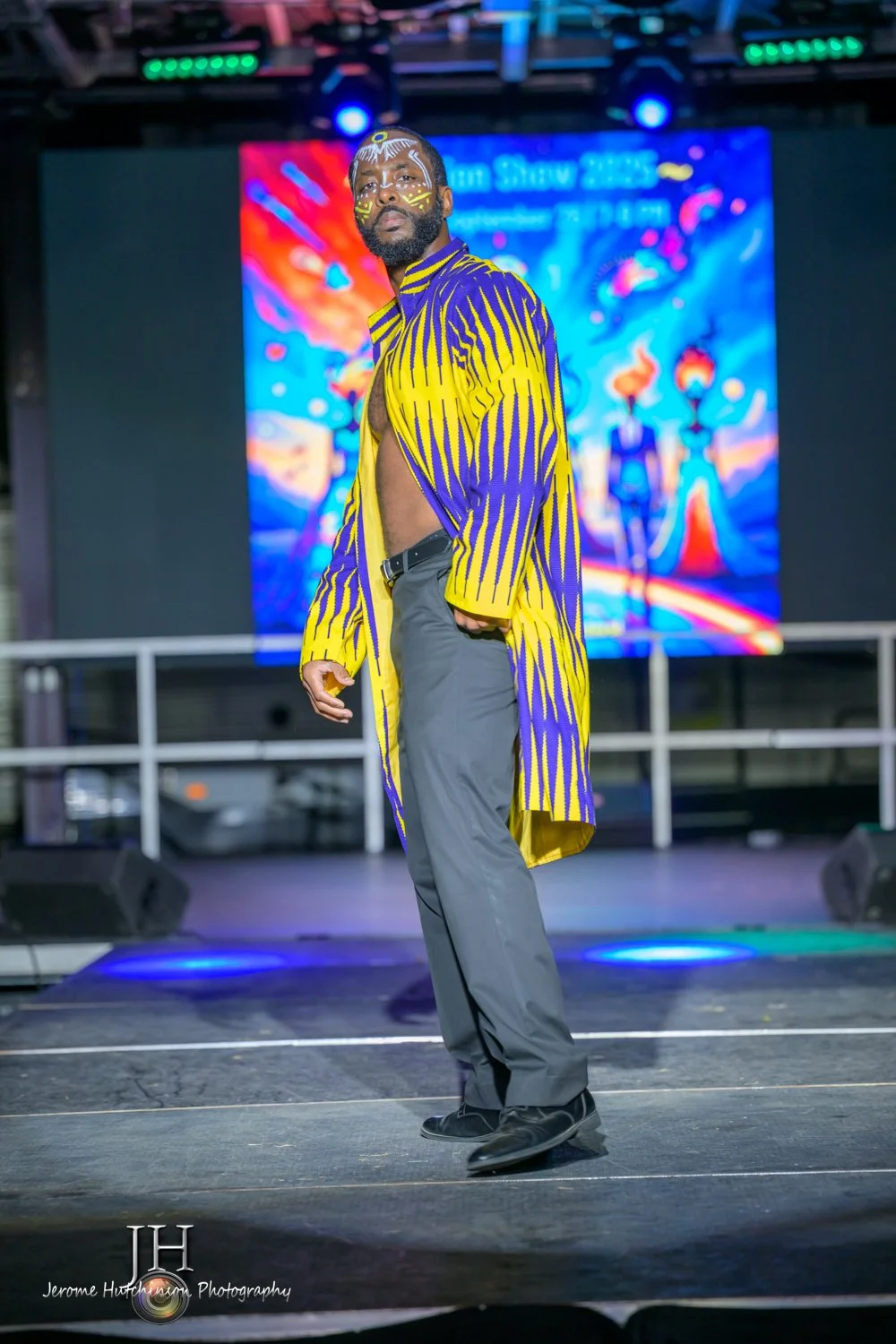 A man with face paint wearing a yellow and purple patterned coat and gray pants stands on stage with a colorful digital background behind him.