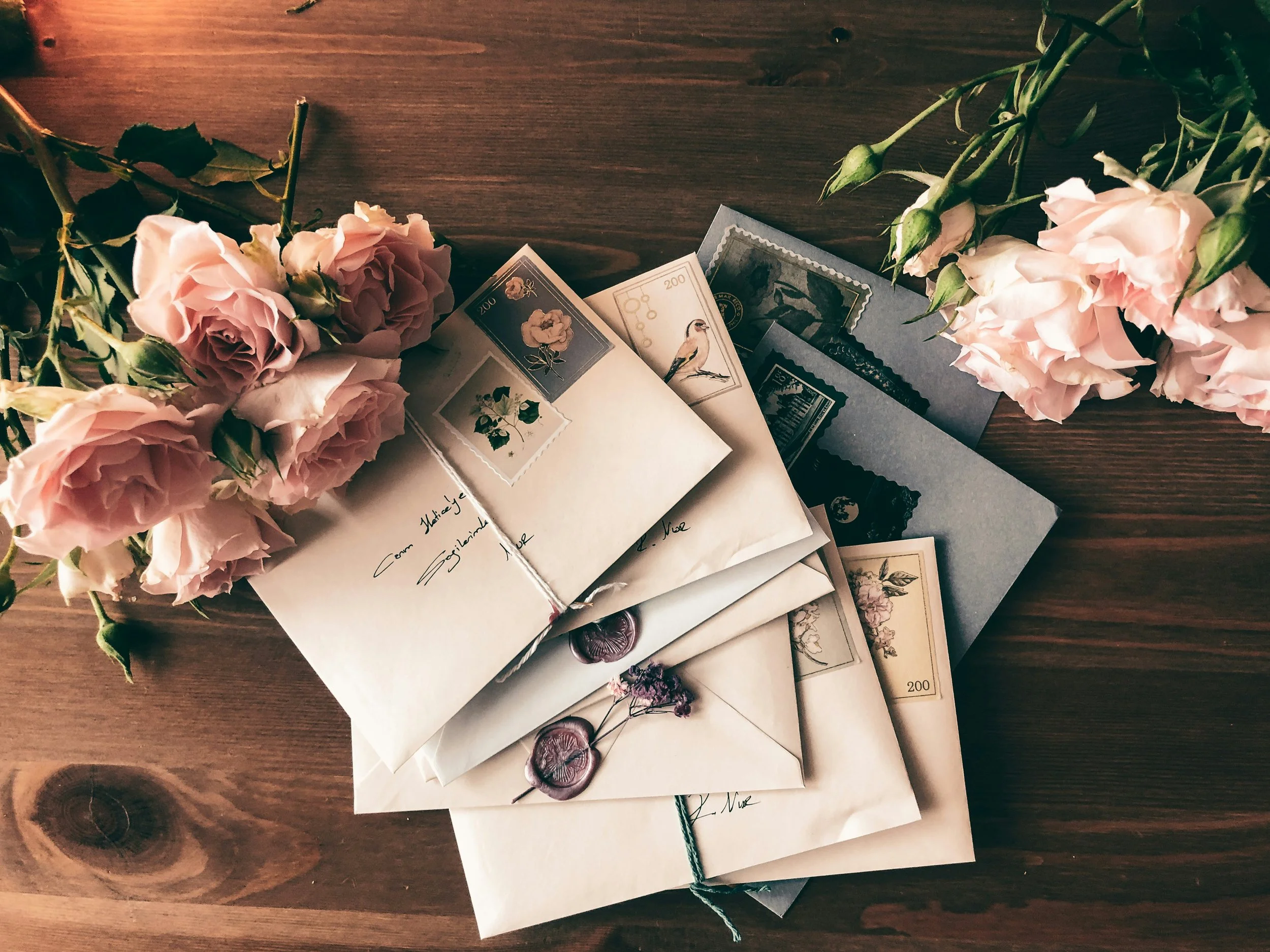 A wooden table with pink roses and several handwritten or printed letters, envelopes, and postage stamps arranged on it.