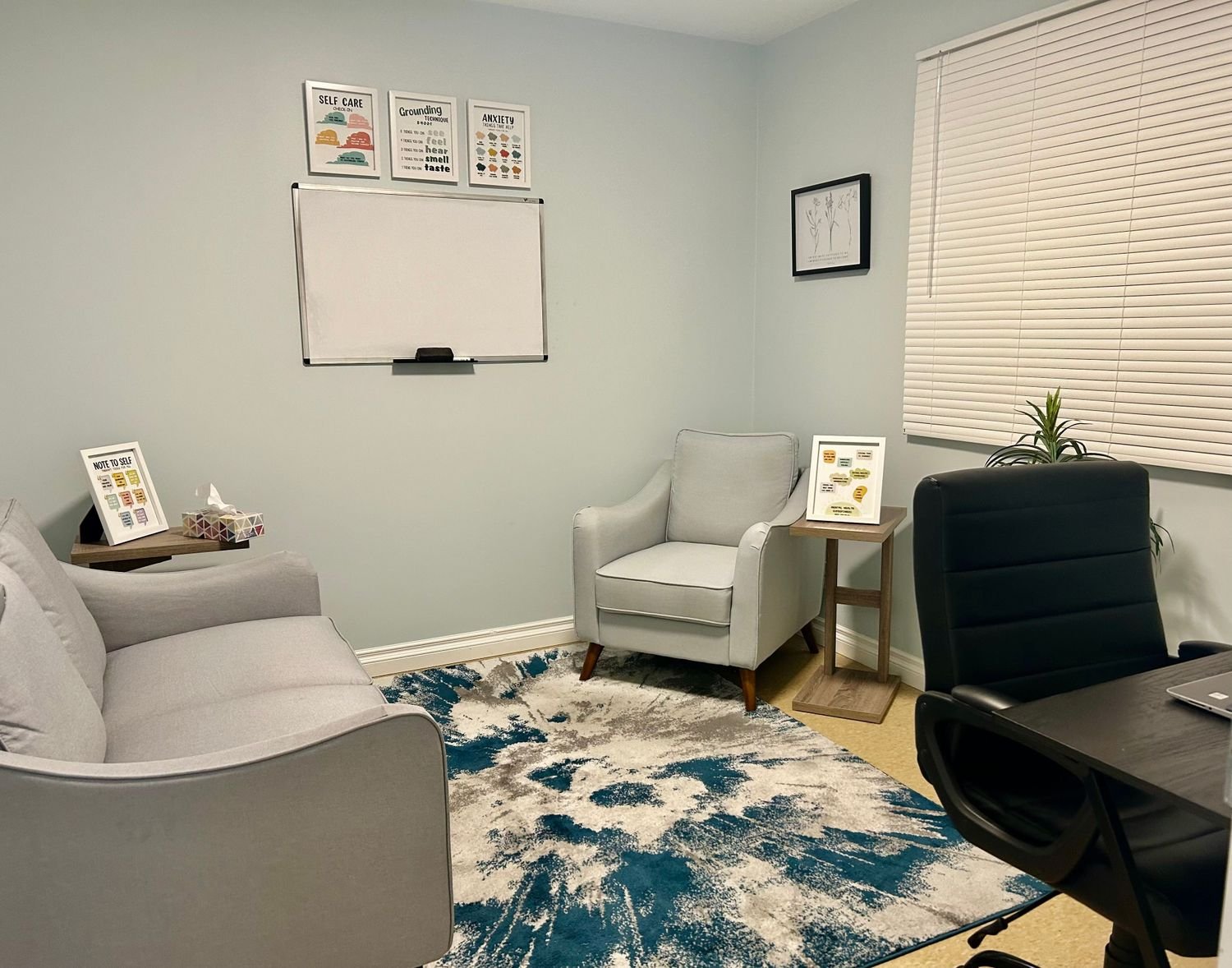 A cozy office with a whitearmchair and a computer conference room setup with a sofa, armchair, picture frames, and a whiteboard on a light blue wall, a window with closed blinds, a small plant, and a patterned rug.