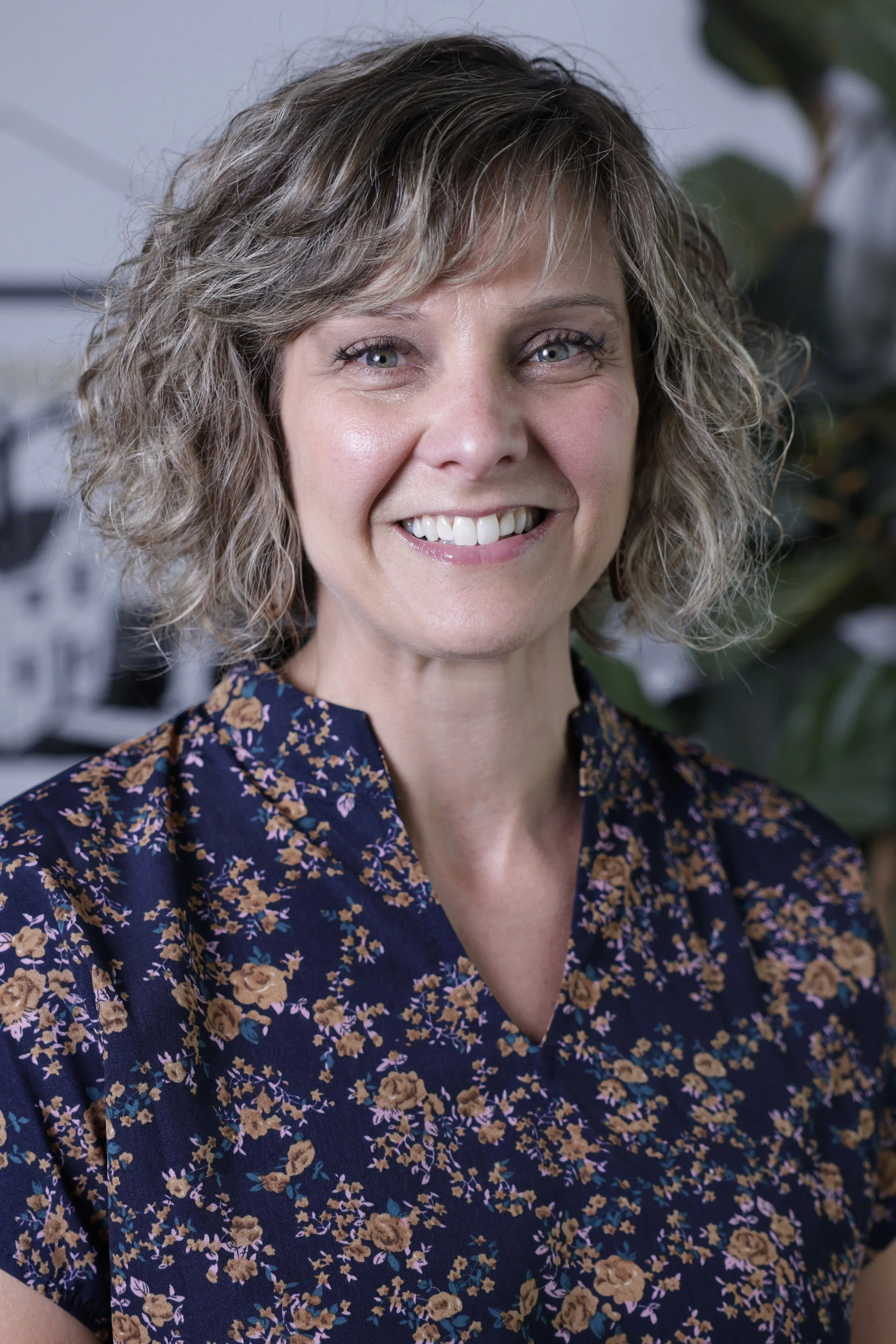 A woman with short, curly blonde hair smiling at the camera, wearing a navy blue blouse with a tan floral pattern, in an indoor setting with green plants in the background.