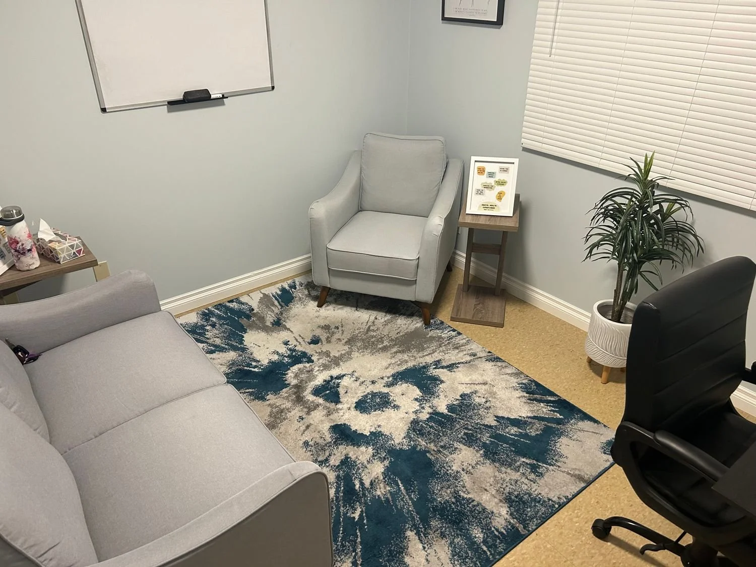 A small office waiting room with a light gray armchair, a black office chair, a gray couch, a blue and gray patterned rug, a small side table with a framed document, a potted plant, and a window with closed blinds.