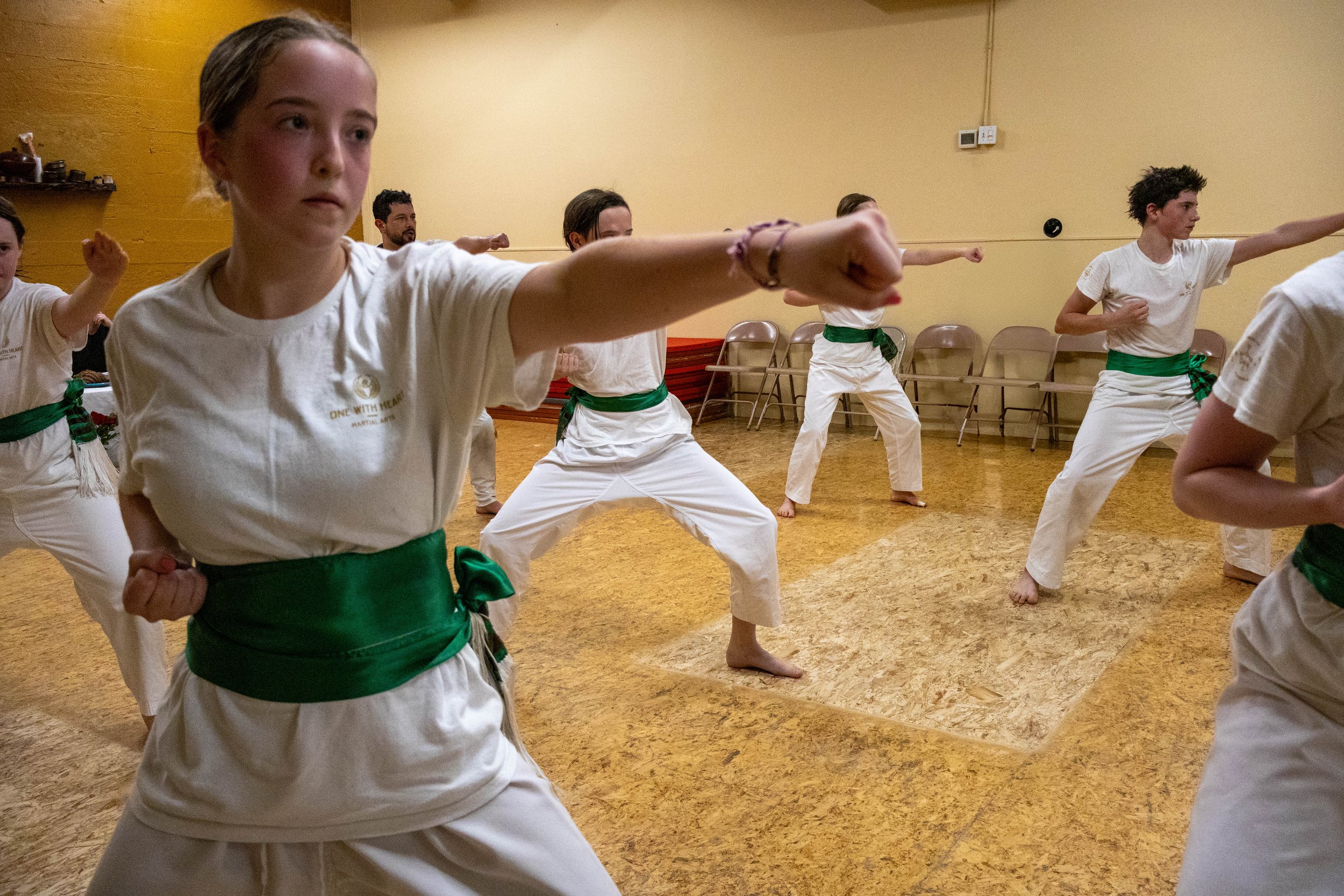 Group of martial arts students practicing in a dojo, all wearing white uniforms with green sashes, performing a punch technique with focused expressions.