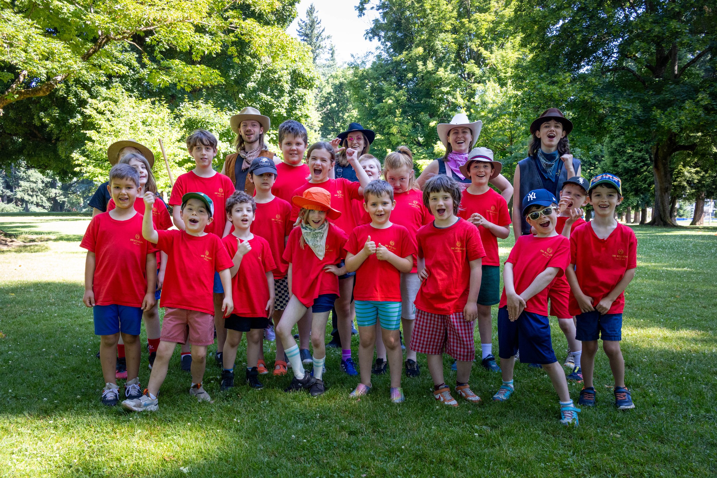 A group of children and a few adults in costumes, some wearing hats, standing outdoors in a park with green trees in the background, smiling and posing for a photo.