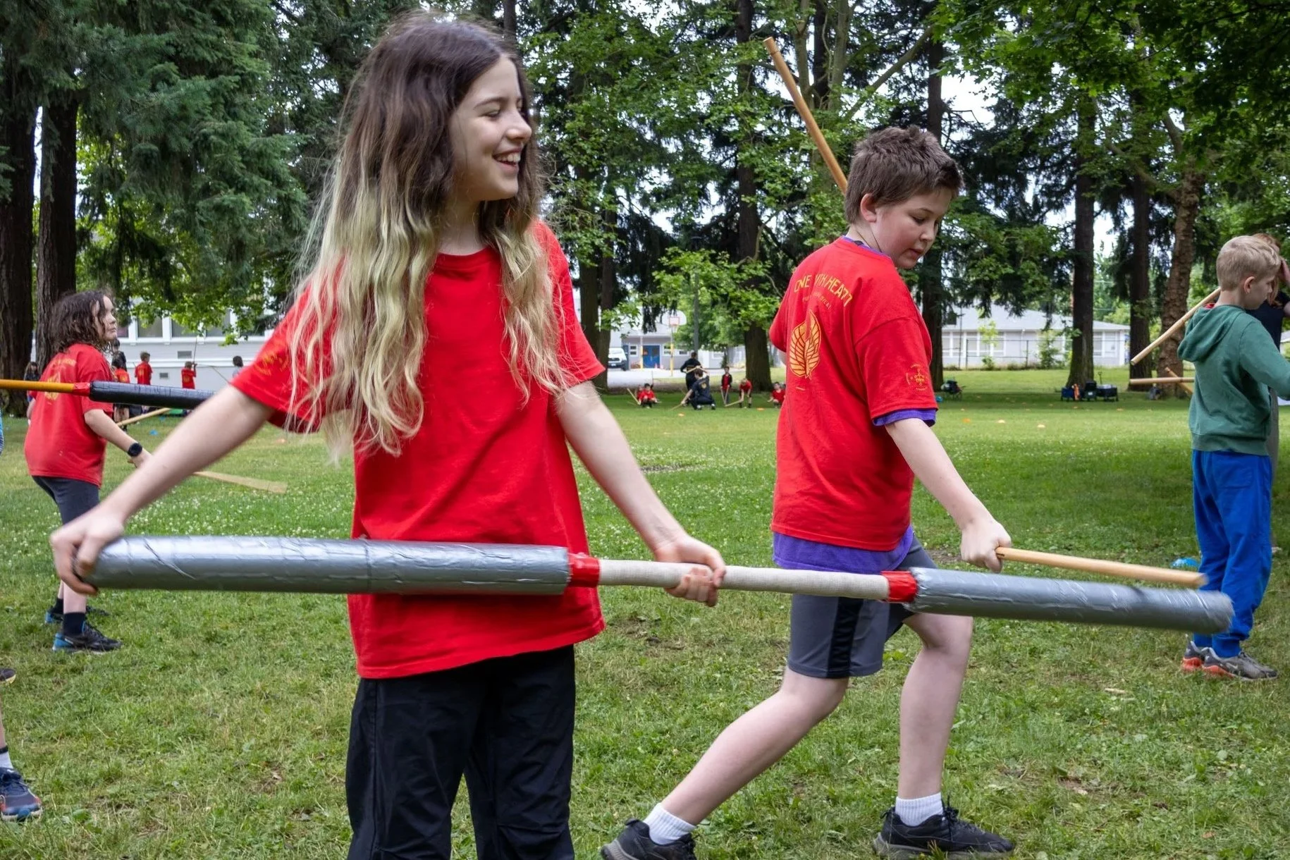 Children participating at an outdoor summer camp with foam swords in a park with green grass and tall trees.