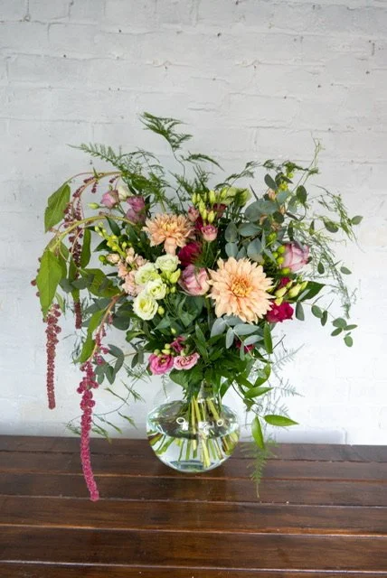 Colorful mixed flower bouquet in a clear glass vase on a wooden table against a white brick wall.