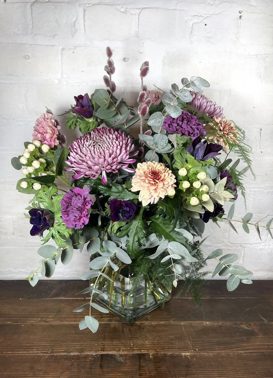 A colorful autumn floral arrangement in a clear glass vase on a wooden surface, featuring chrysanthemums, roses, berries, and various fall leaves.