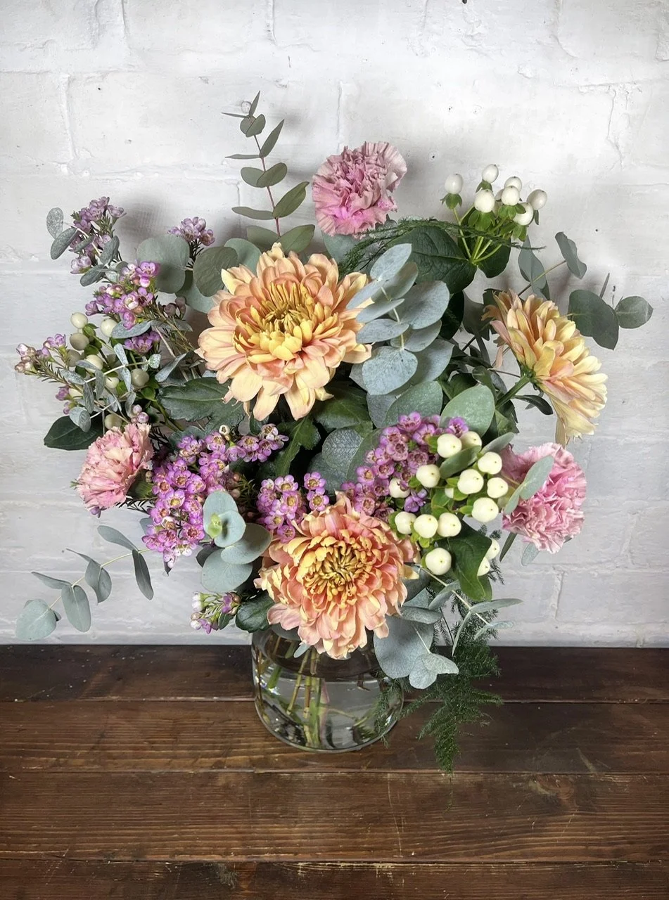 A romantic, wreath decorated with pink, purple and red flowers, dried foliage, cinnamon sticks, and hanging velvet ribbons at the base, hung on white wall.