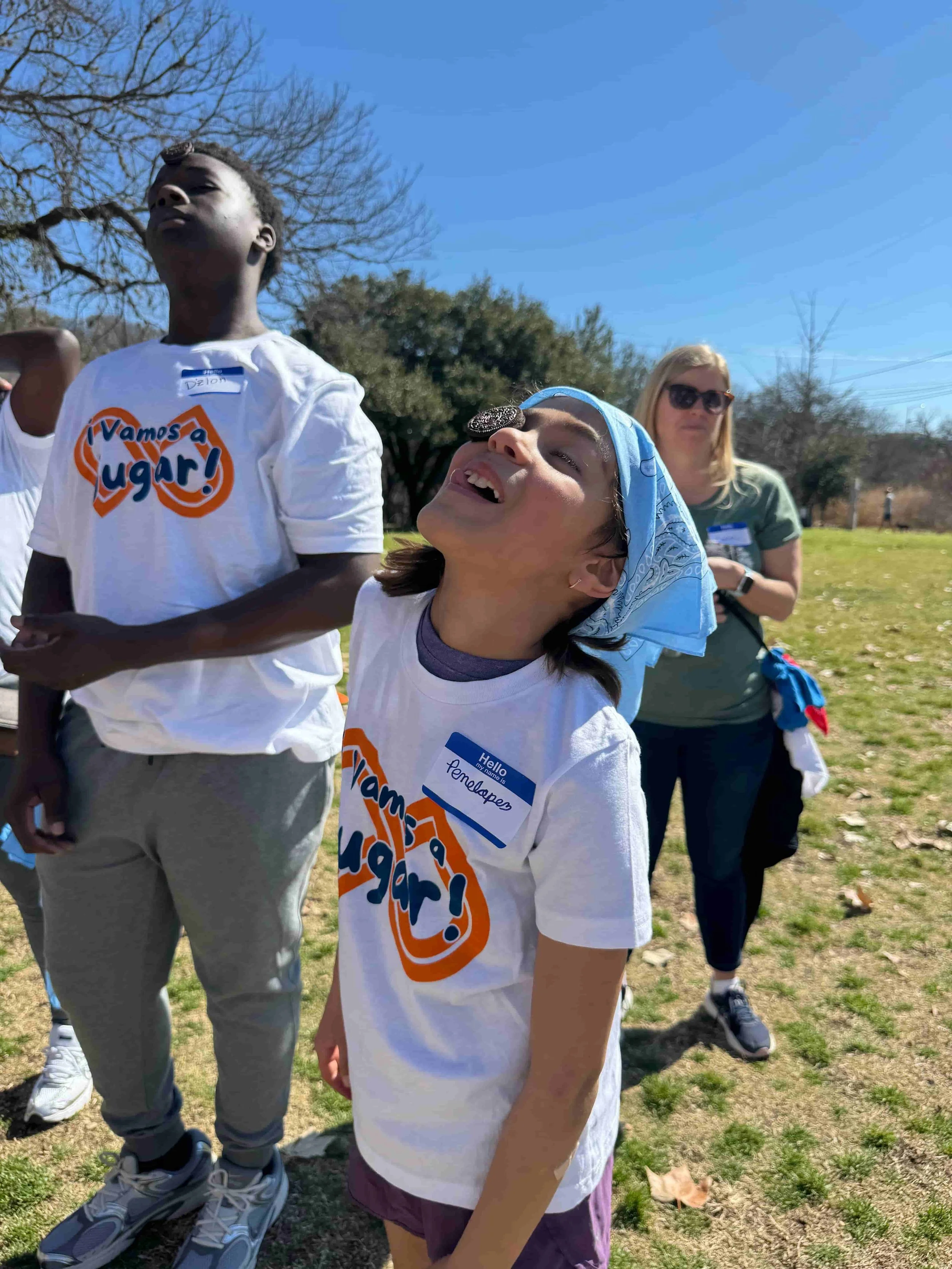 Two kids in Throwback "¡Vamos a Jugar!" t-shirts laugh and look up mid-game during a field day event