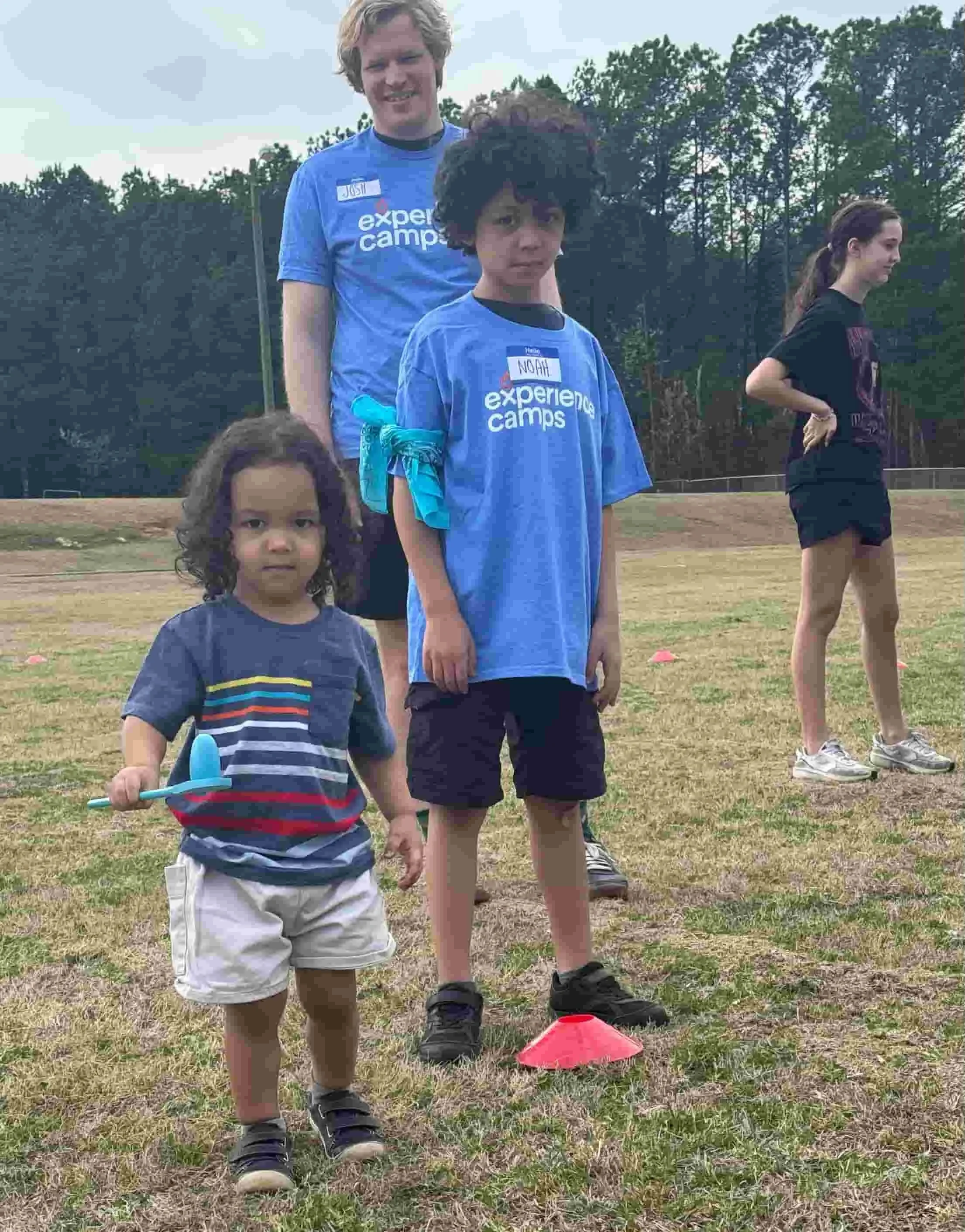 Two young campers in blue Experience Camps t-shirts stand on a sports field next to a Throwback volunteer counselor